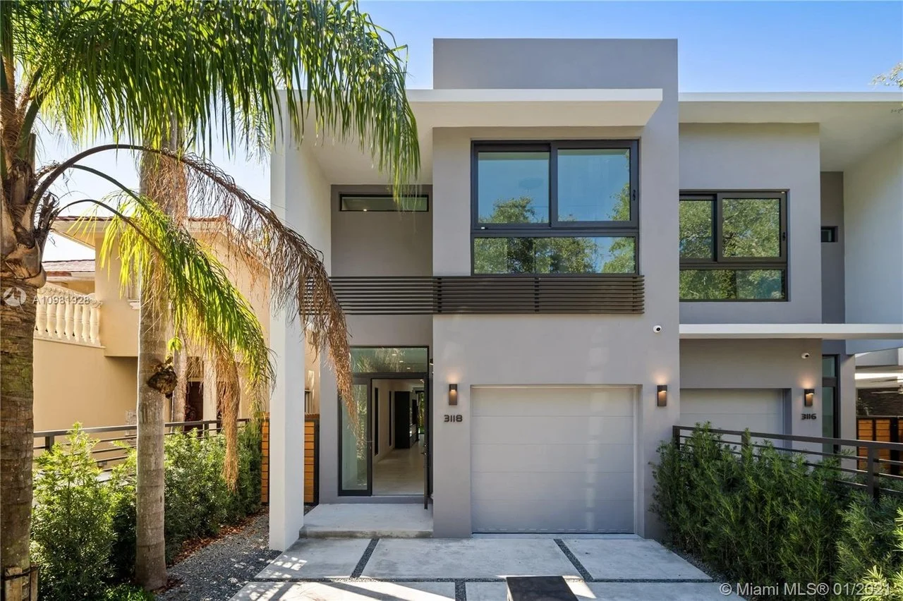 Modern two-story house with white exterior, large windows, and a flat roof. The front entry is next to a closed garage door, illuminated by wall-mounted lights. There are palm trees and shrubs along the pathway leading to the front door, under a partly sunny sky.