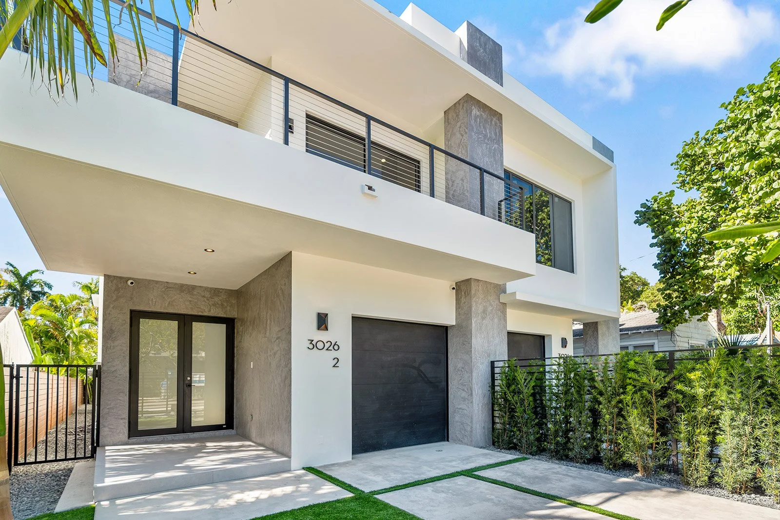 Modern multi-story house with white and gray exterior, black accents, glass doors, and a small front yard with concrete driveway and green grass