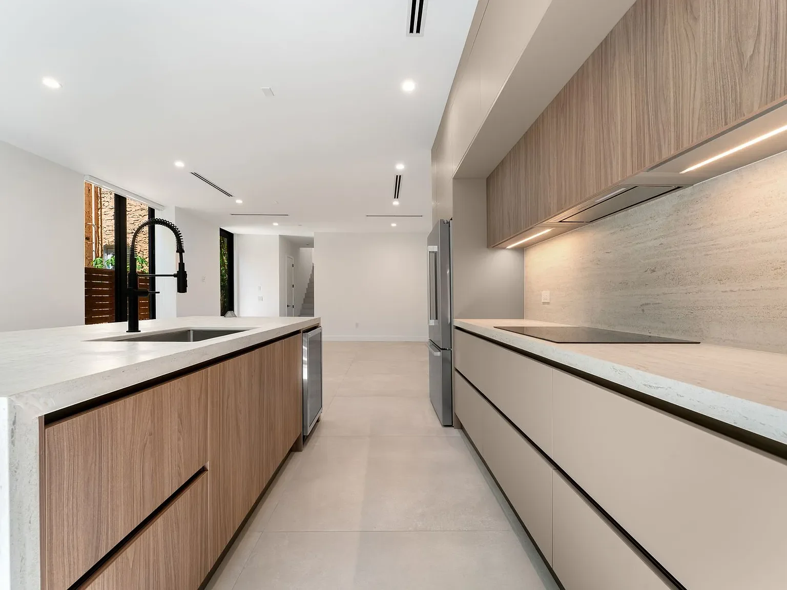 Modern kitchen with minimalist design, featuring a large white island with a black faucet, wood and cream-colored cabinetry, built-in appliances, and bright lighting.