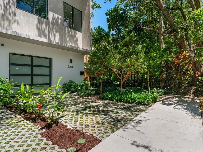 Modern house with a concrete driveway, lush green plants, and trees in a sunny, landscaped yard.