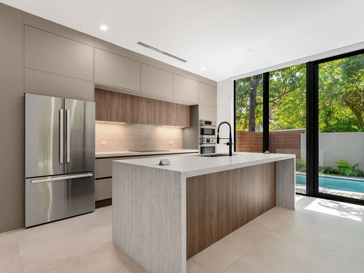 Modern kitchen with beige cabinets, a white marble island, stainless steel refrigerator, built-in oven and microwave, black faucet, and large sliding glass doors leading to a backyard with trees and a pool.