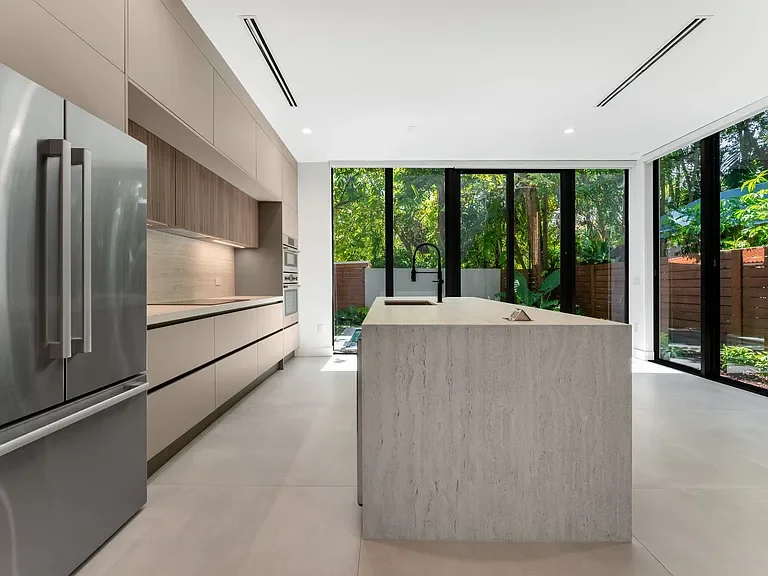 Modern kitchen with beige cabinets, stainless steel refrigerator, marble island, and large glass sliding doors leading to a garden.