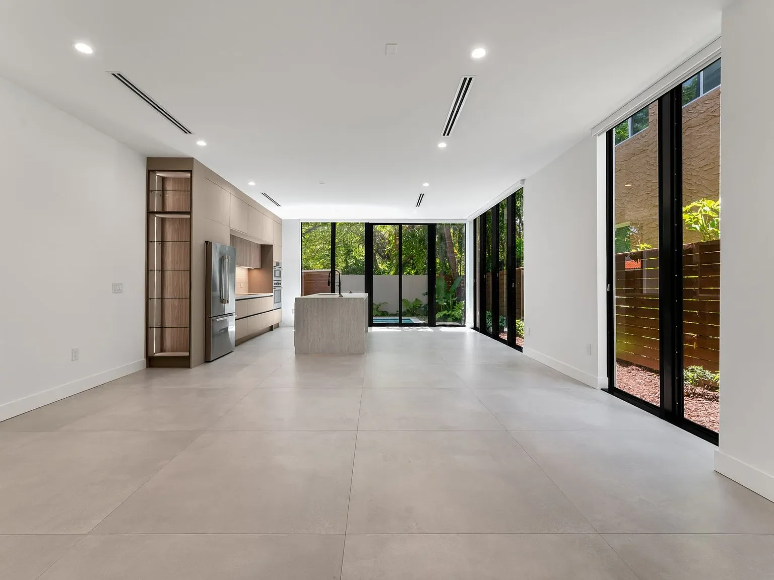 Empty modern kitchen with large windows, white walls, beige floor tiles, and built-in cabinetry.