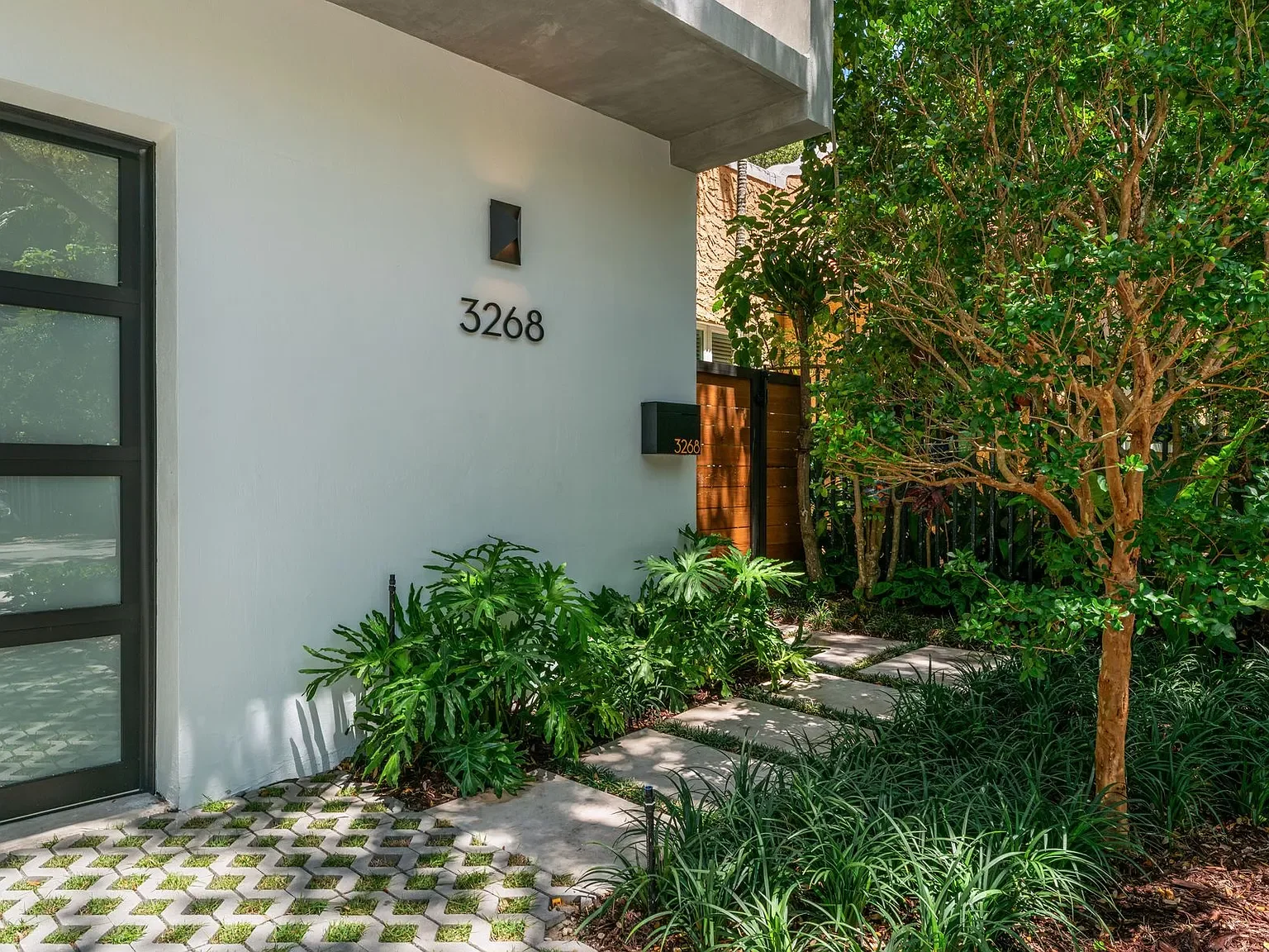 Modern house entrance with the number 3268 on the wall, surrounded by lush green plants, trees, and a paved pathway.