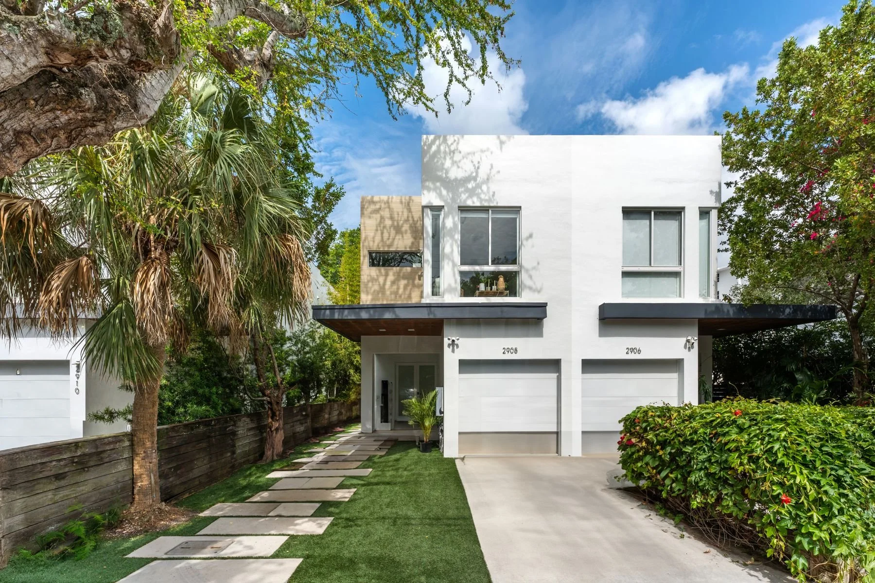 Modern white two-story house with a flat roof, large windows, and attached garage, surrounded by trees and greenery, with a stepping stone pathway and a well-manicured lawn.