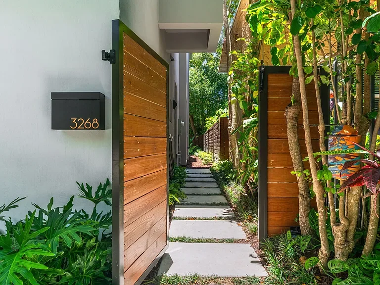 A modern house entrance with a wooden gate, house number 3268, a concrete pathway, and lush green plants and trees along the side.
