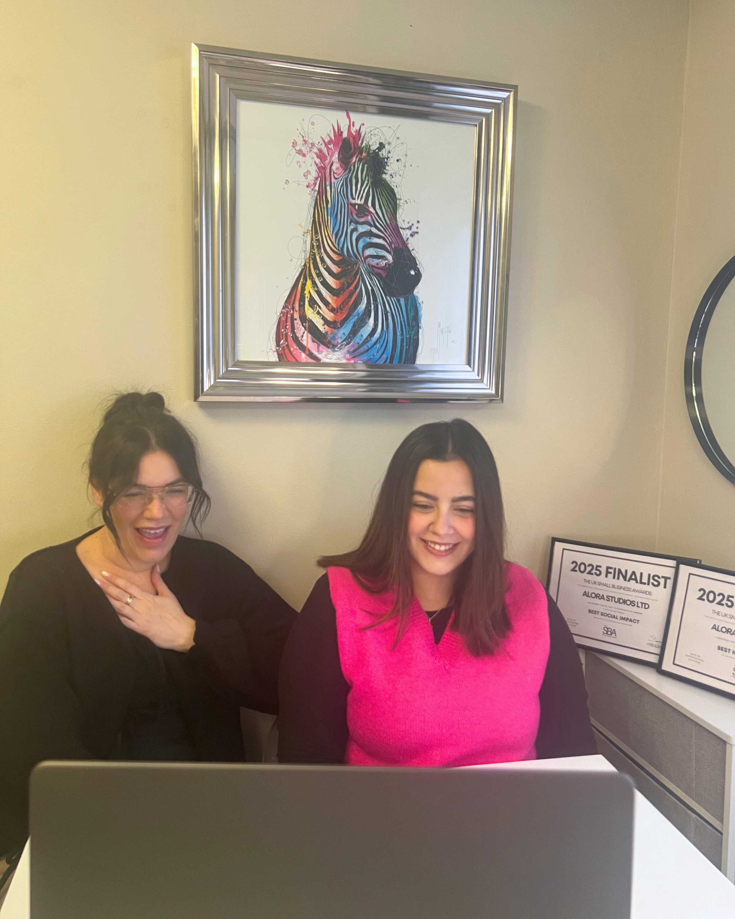 Two women sitting at a desk smiling at a laptop, with colorful awards on the wall and a framed picture of a zebra above them.