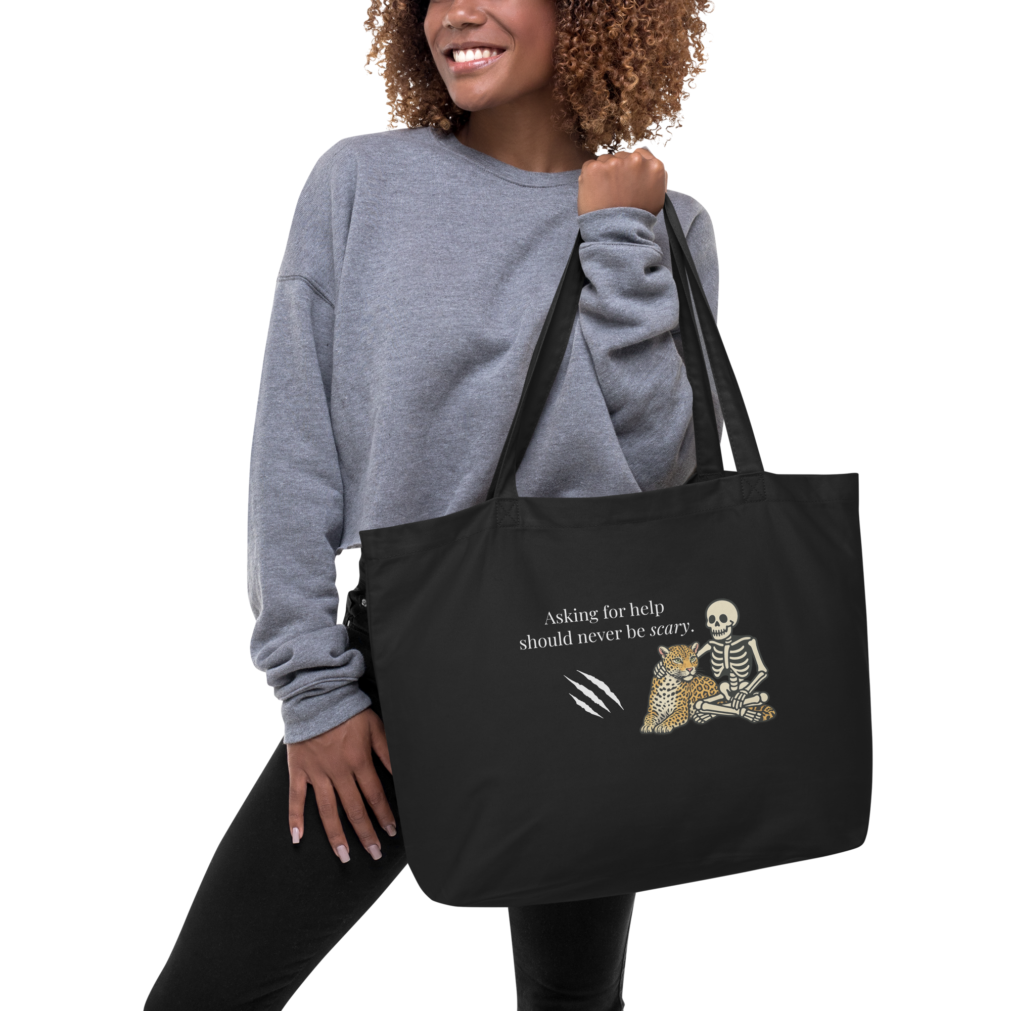 A woman wearing a gray sweatshirt holding a black tote bag with the text 'Asking for help should never be scary.' and a graphic of a skeleton, a leopard, and claw marks.