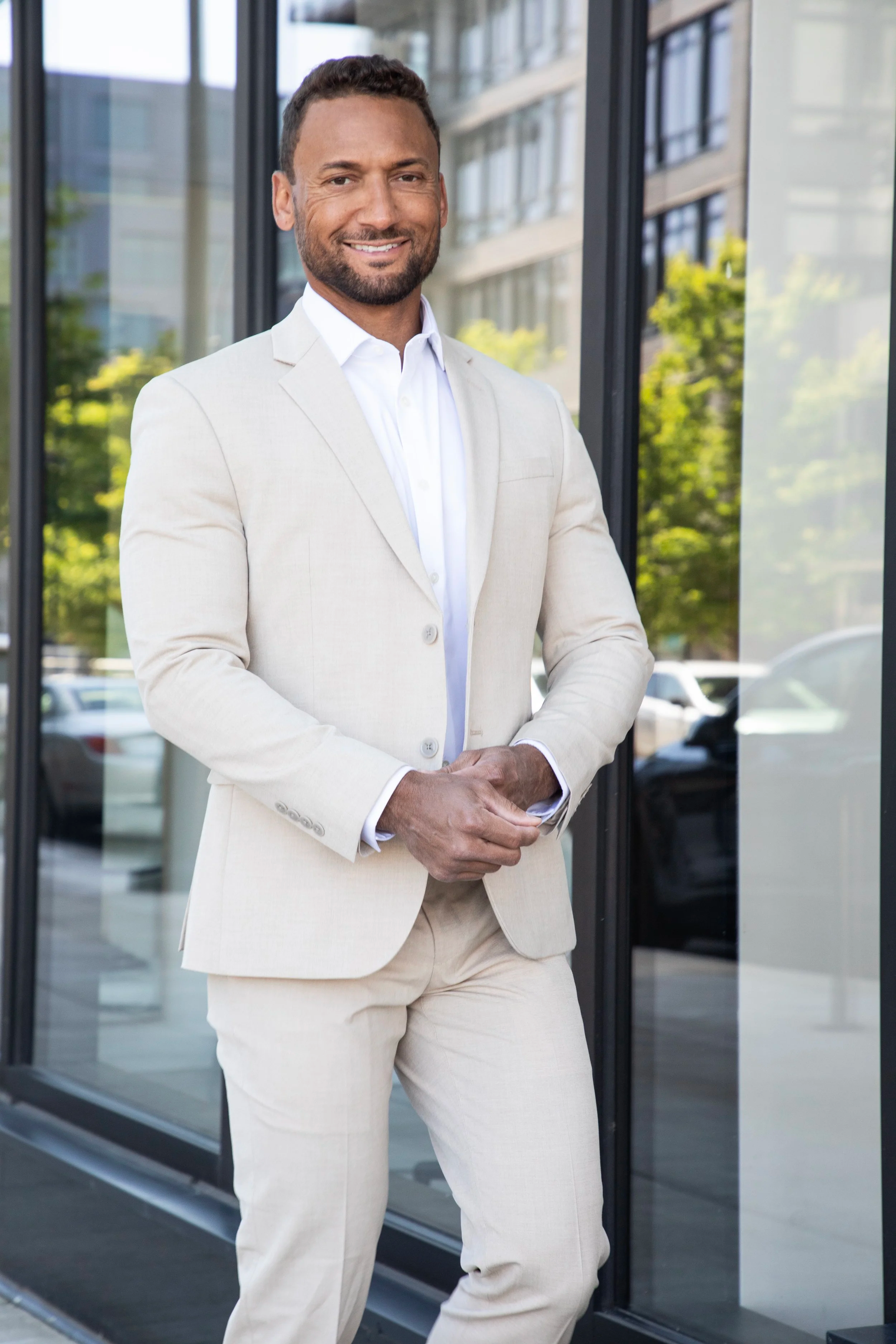 Man in cream-colored suit standing outside building with glass windows, smiling with arms crossed.