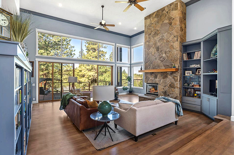 Spacious living room with large windows showing trees outside, a stone fireplace, blue built-in shelves, and modern furniture including two sofas and a round side table with a green vase.