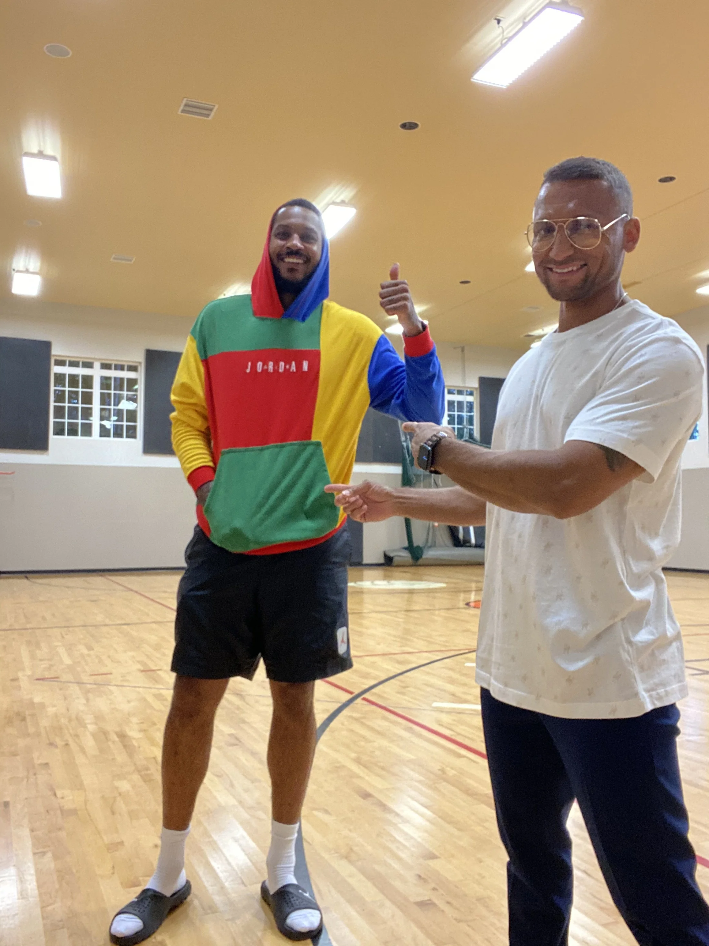 Two smiling men in a gymnasium, one wearing a colorful hoodie and sandals with socks, the other wearing glasses and a white t-shirt, are pointing at each other.
