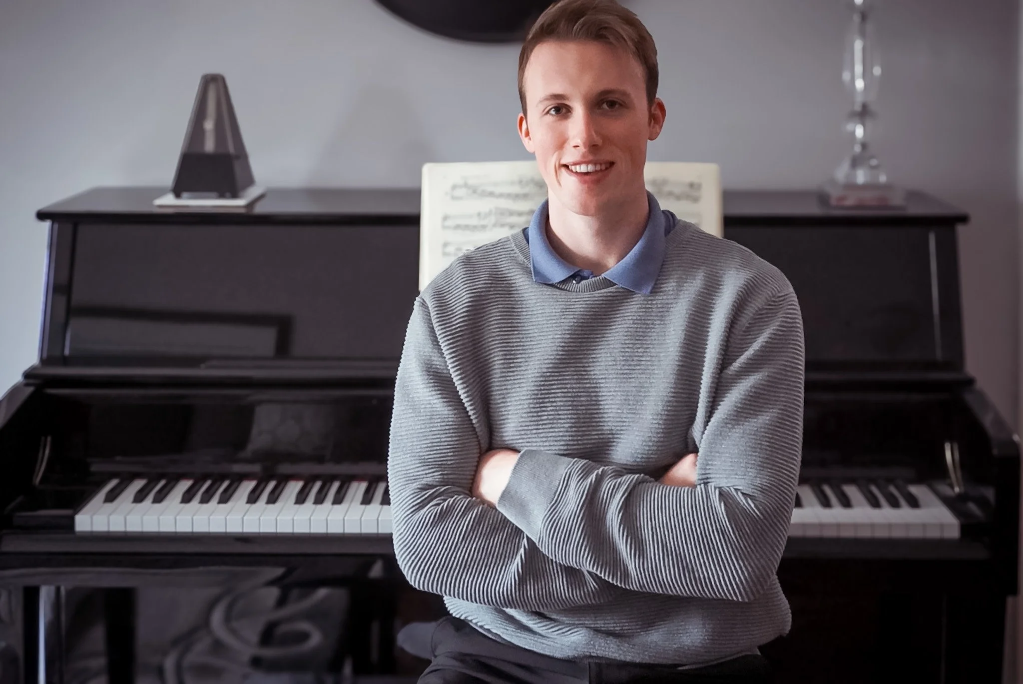 A young man with short brown hair, wearing a gray striped sweater over a blue collared shirt, sitting with arms crossed in front of a black upright piano with sheet music and a metronome on top.