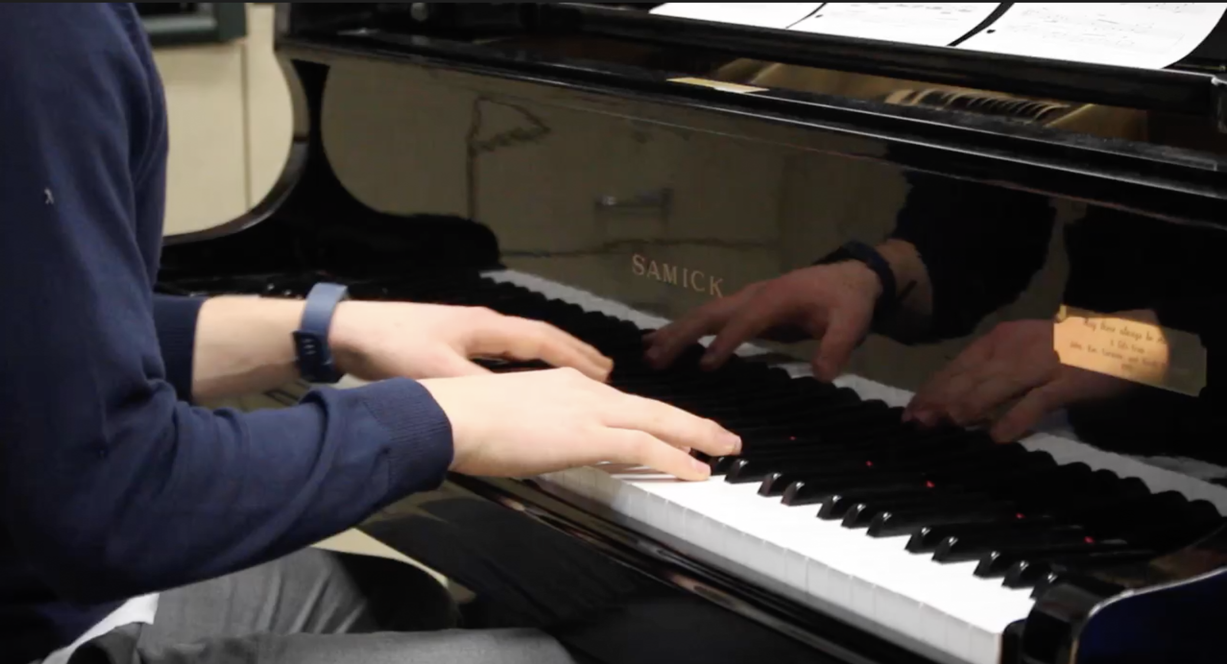 Person playing a black grand piano. The person's hands are on the keys, and their reflection is seen on the polished surface of the piano.