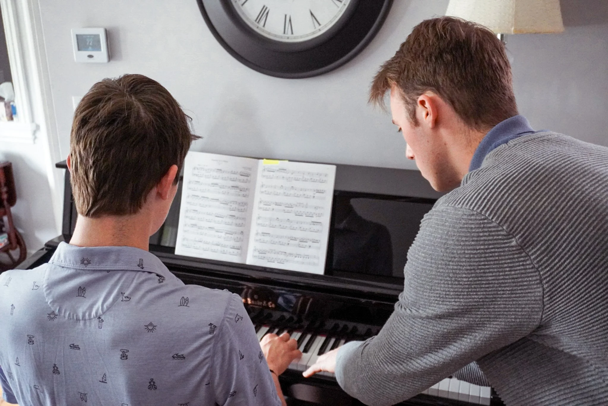 A music teacher is teaching one of his students. They are in front of a black upright piano. The teacher is showing proper hand placement to the student. The student will begin to play the music in front of him