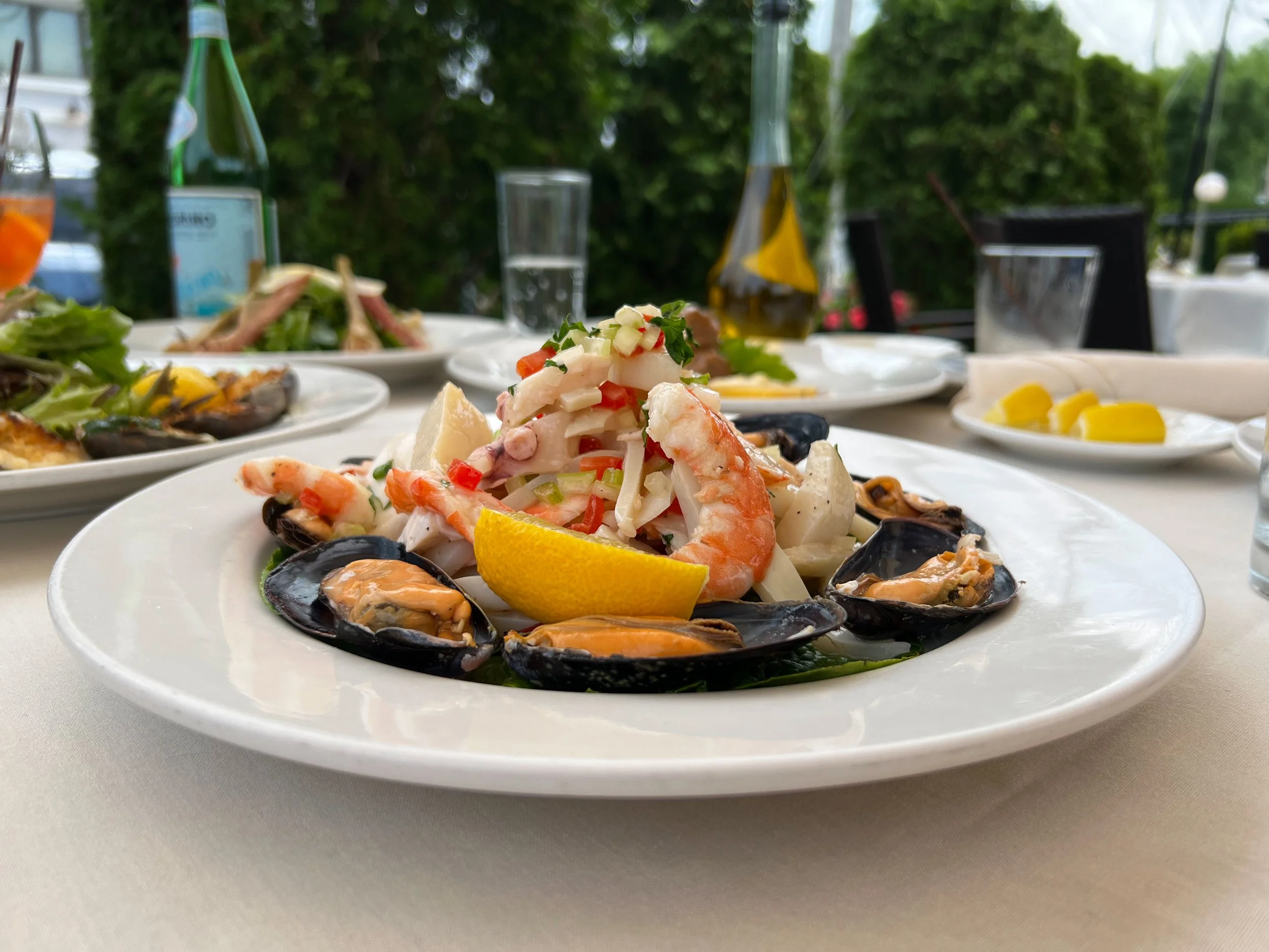 Seafood salad with shrimp, mussels, squid, and vegetables garnished with lemon wedge on a white plate, set outdoors with additional dishes and drinks in the background.