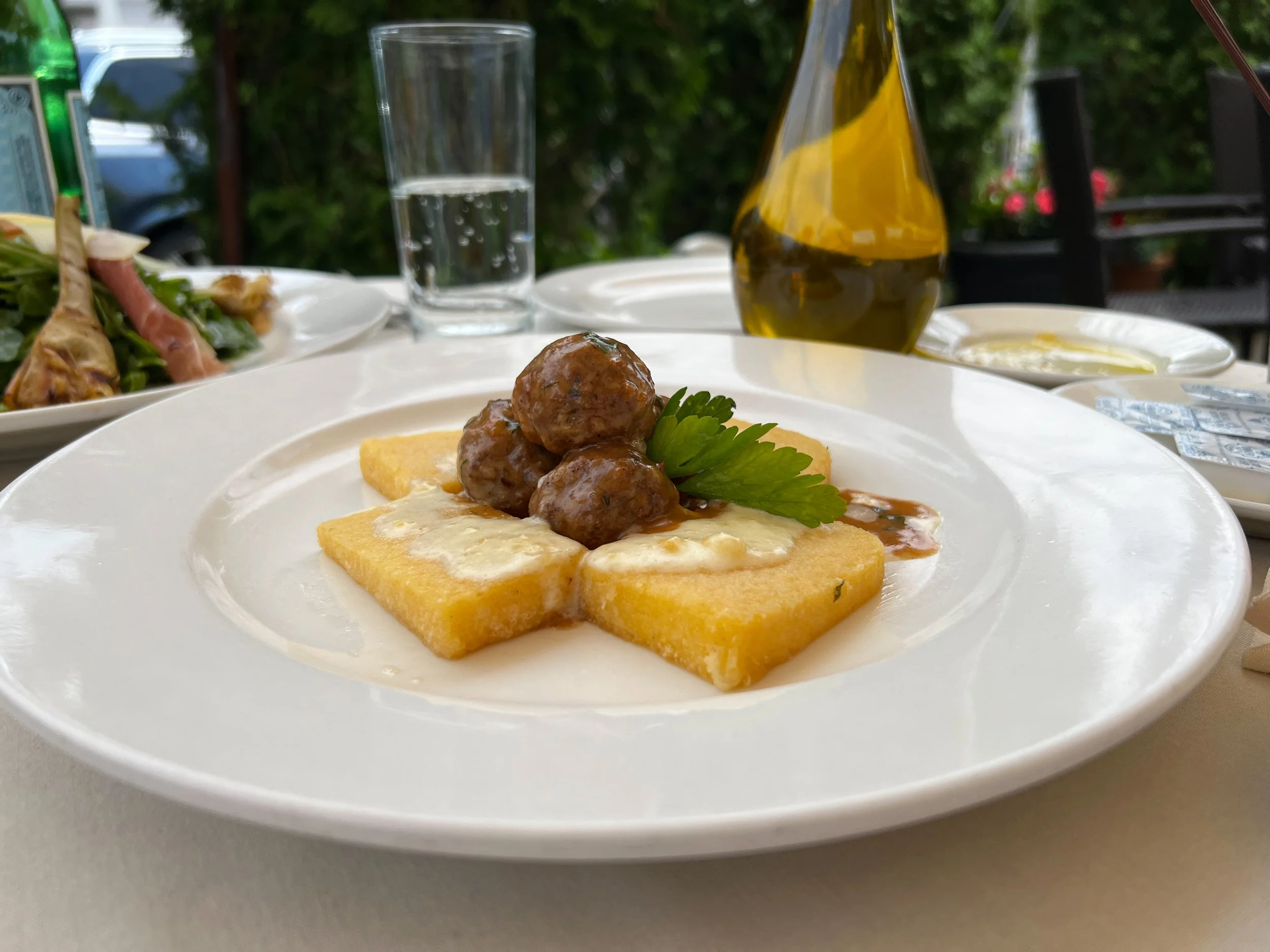 A white plate with fried polenta cubes topped with meatballs in sauce and garnished with a sprig of cilantro, set on an outdoor dining table.