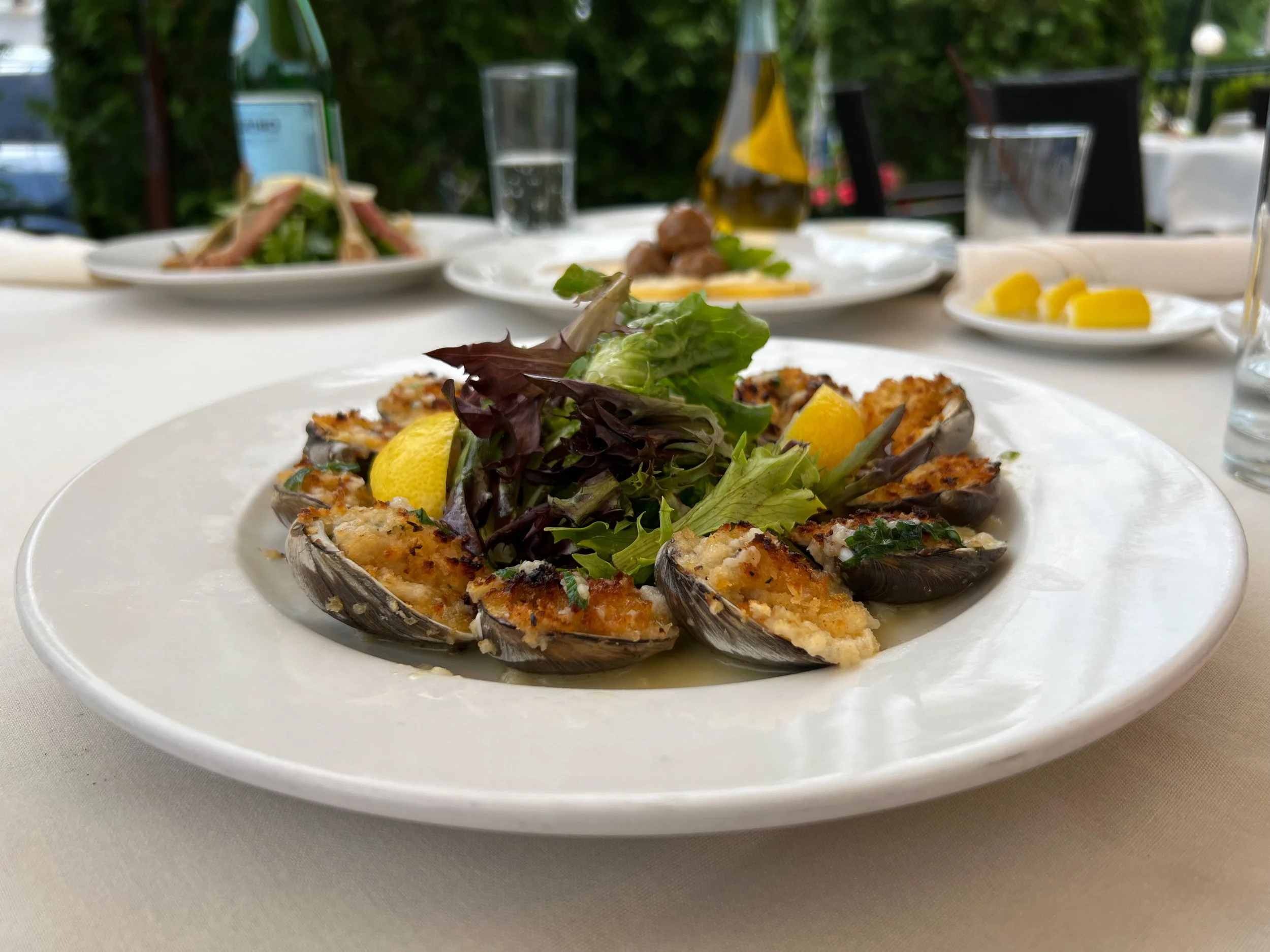 A white plate of grilled stuffed mussels garnished with lemon wedges and mixed greens on a table at an outdoor restaurant.