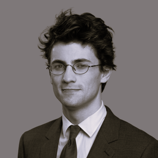 Portrait d'un jeune homme portant des lunettes, chemise blanche et veste de costume, avec des cheveux bouclés.