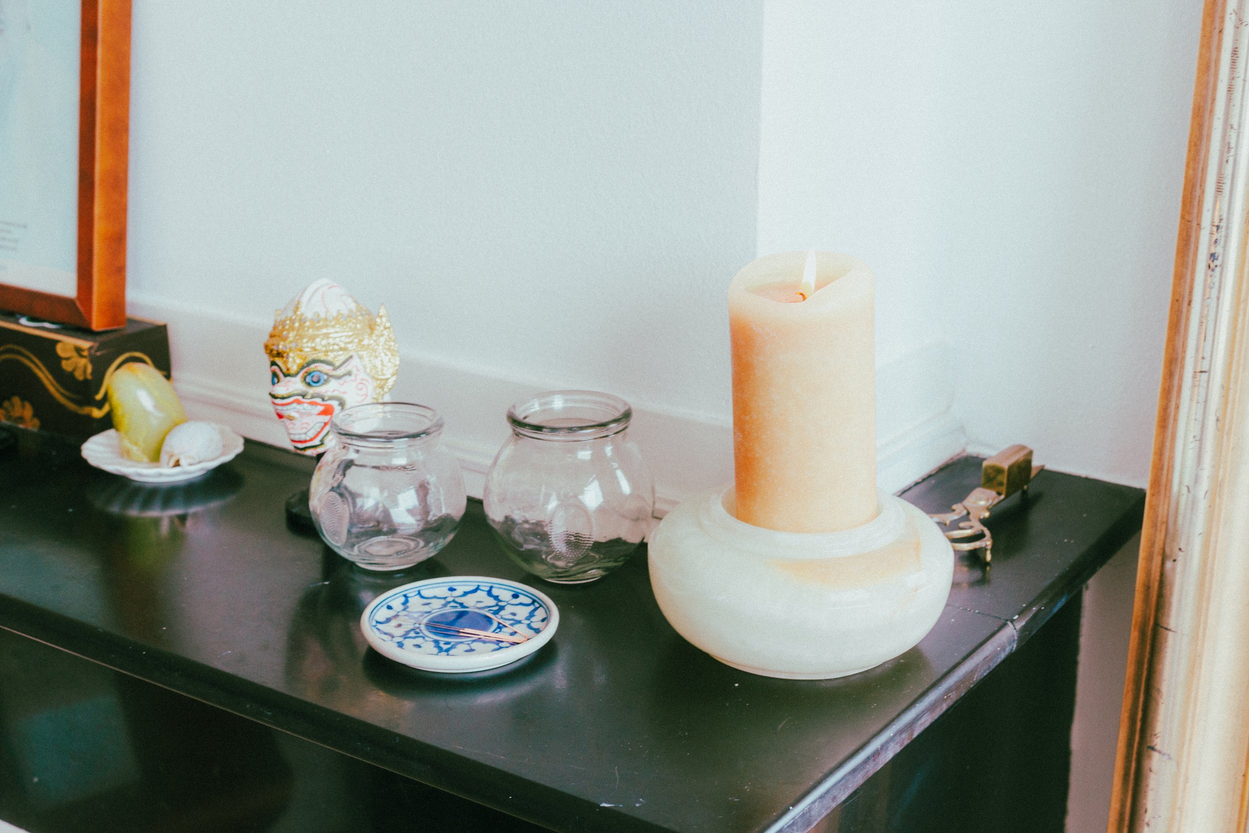 Decoratieve voorwerpen op een zwarte tafel, waaronder een grote kaars, een masker, twee glazen potjes, een klein bord met een naald.