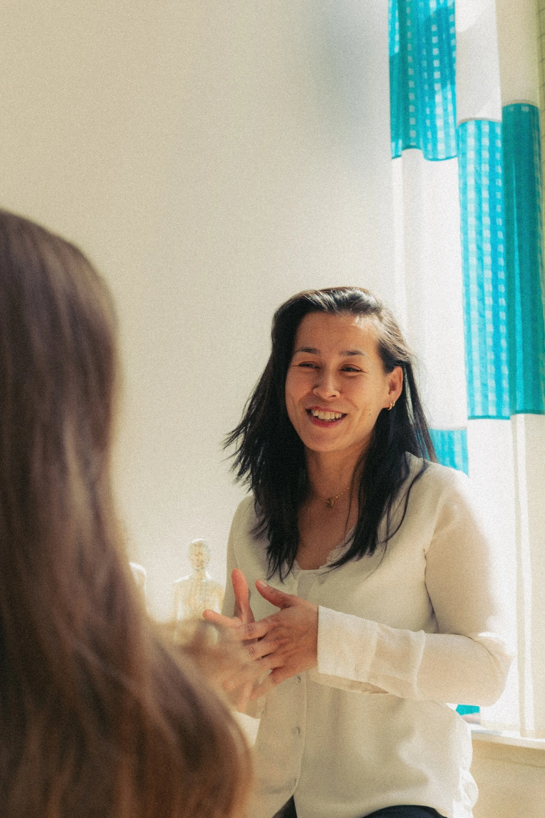 Twee vrouwen die in een kamer praten, een glimlachende vrouw in een witte blouse staat bij een raam met blauwe en witte gordijnen.