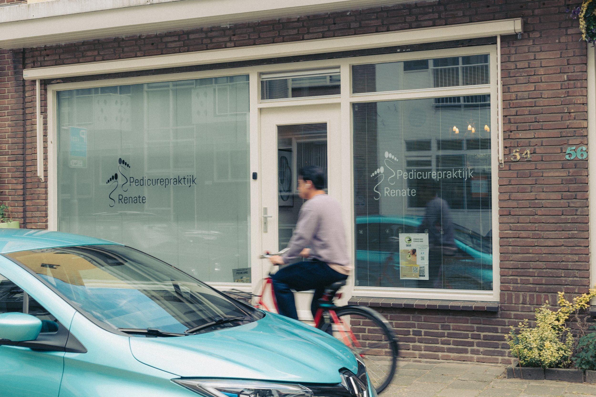 Een man fietst voor een winkel met ramen die de tekst 'Pedicurepraktijk Renate' dragen. Een lichtblauwe auto staat geparkeerd op de straat. De winkel zit in een gebouw met een bakstenen gevel, en het raam heeft horizontale blinds.