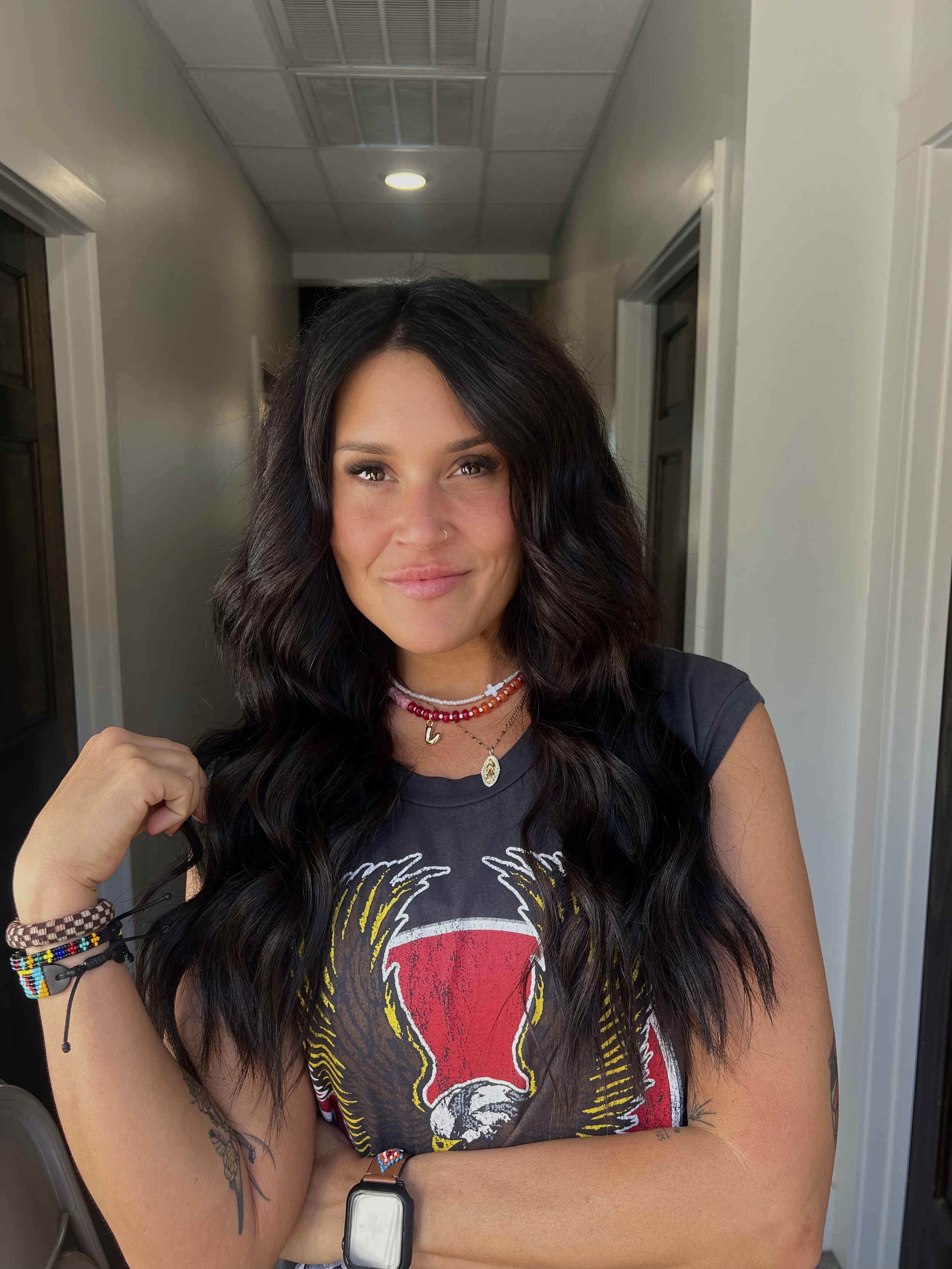 Woman with long dark wavy hair wearing a black t-shirt with a graphic design, multiple necklaces, bracelets, and a smartwatch, standing in a hallway.
