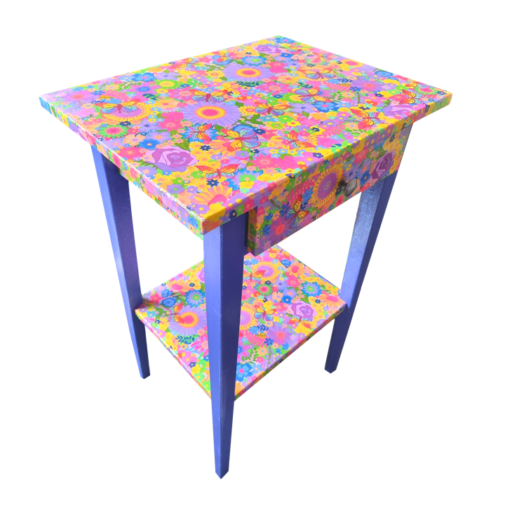 Colourful small table with floral and butterfly patterns on the top, drawer, and lower shelf, with blue legs.