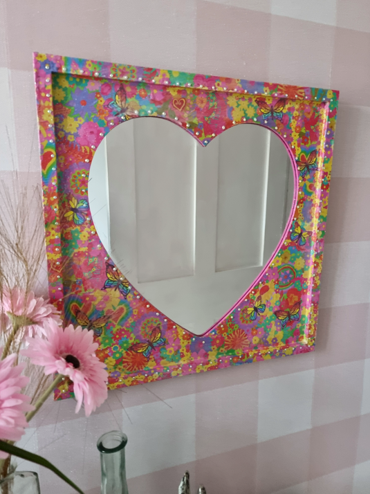 Decorative square mirror with a heart-shaped reflection area, surrounded by a Colourful, floral, and butterfly pattern frame, mounted on a pink and white checkered wall, with pink flowers and a glass bottle in the foreground.