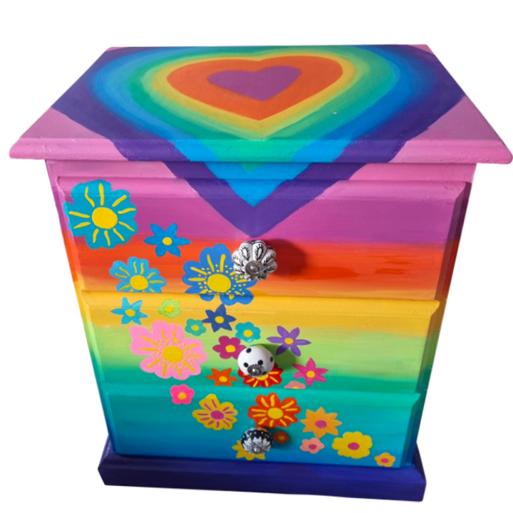 A Colourful chest of bedside drawers painted with rainbow stripes, decorated with various flower stickers, and topped with a rainbow heart design.