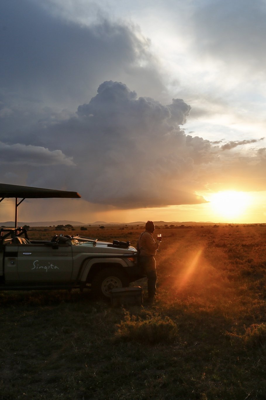 Person standing next to an off-road vehicle called Singita, holding a drink during sunset in a vast open plain with a dramatic sky and clouds.