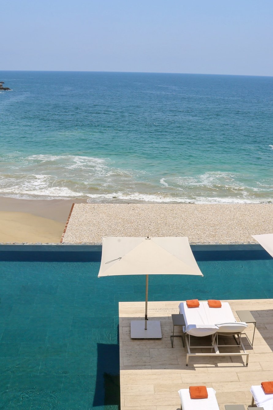 View of a swimming pool overlooking the beach and ocean, with lounge chairs, an umbrella, and towels on the poolside.