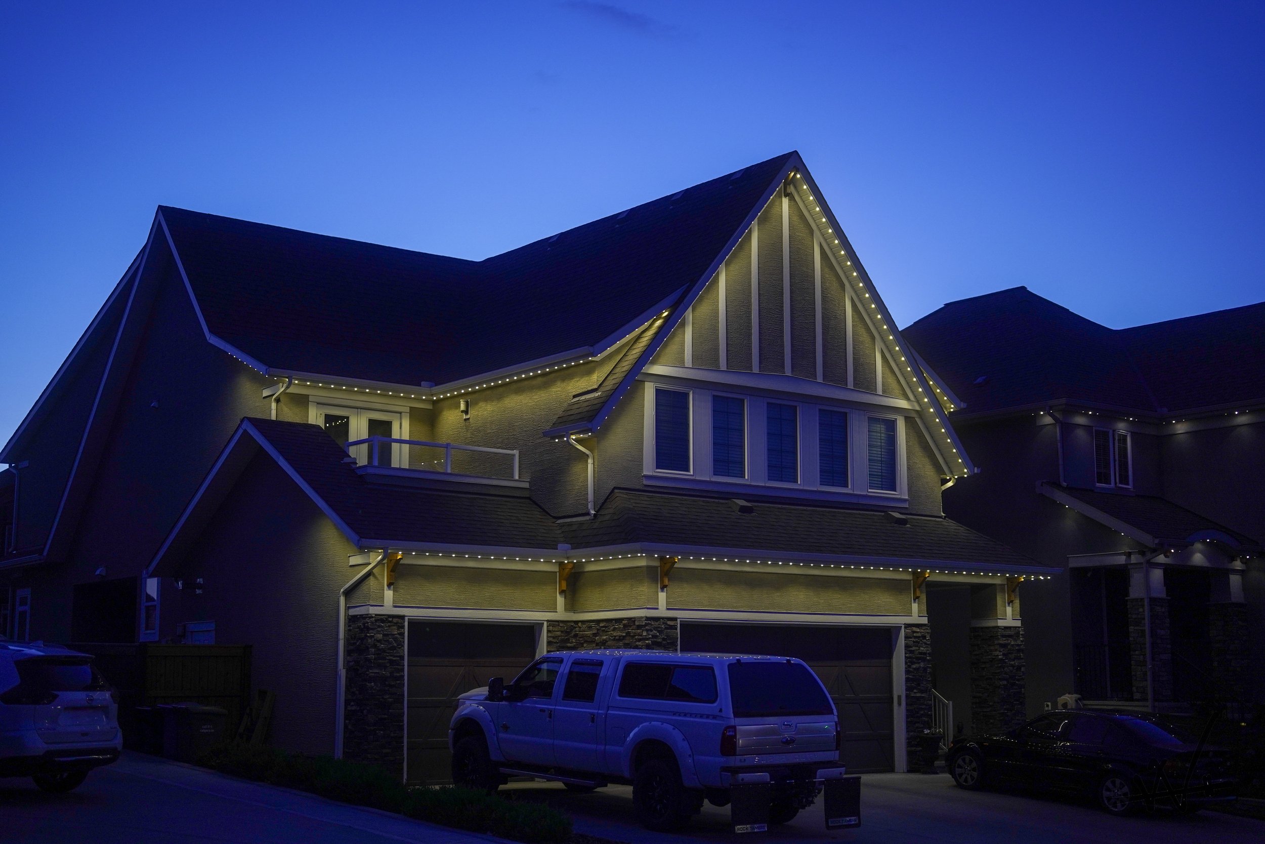 A two-story house at dusk illuminated with permanent track lighting outlining the roof and balconies, with parked cars in the driveway.
