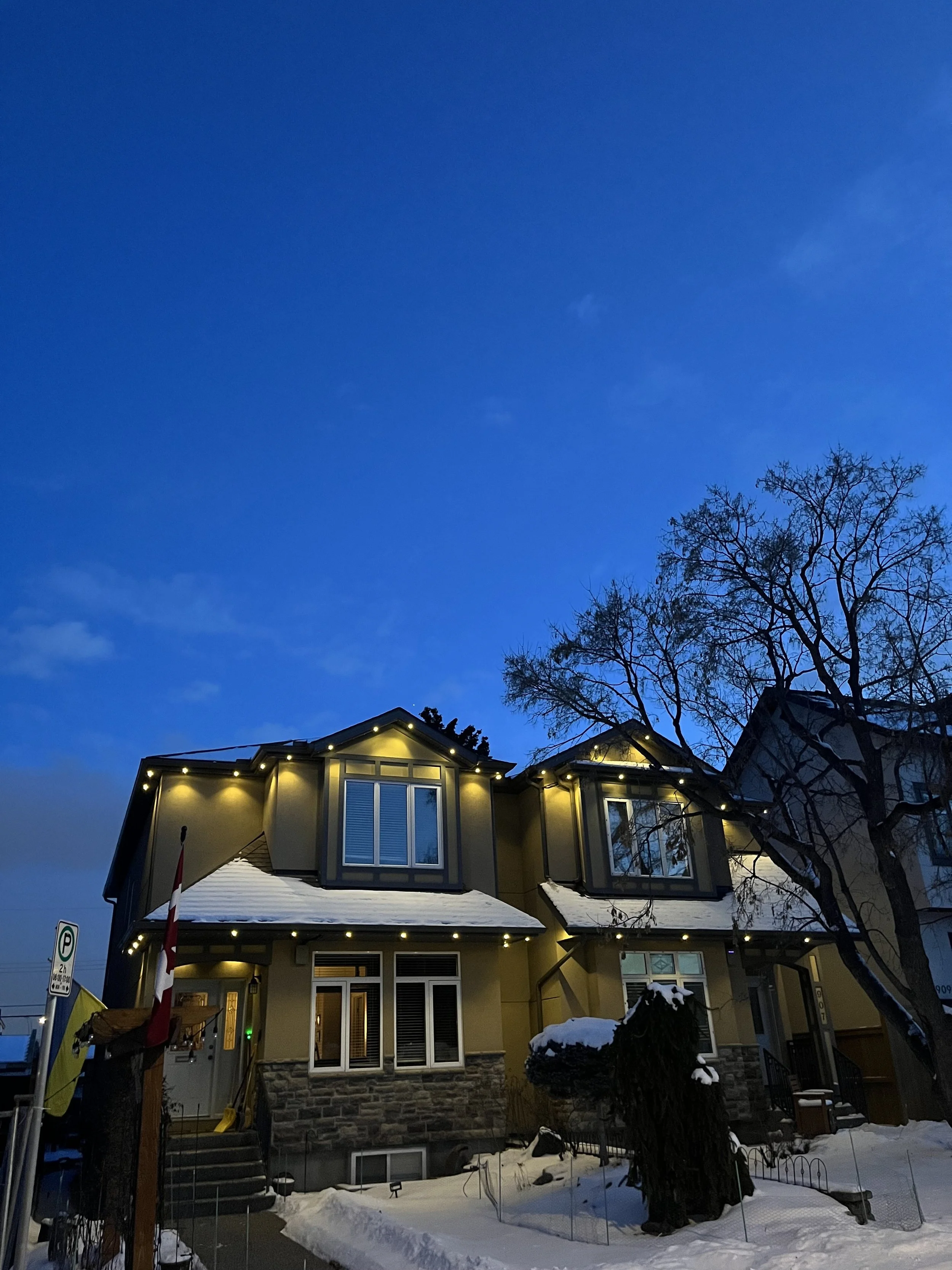 A two-story house decorated with permanent track lighting during winter, with snow on the roof and ground, a leafless tree, and a clear blue evening sky.