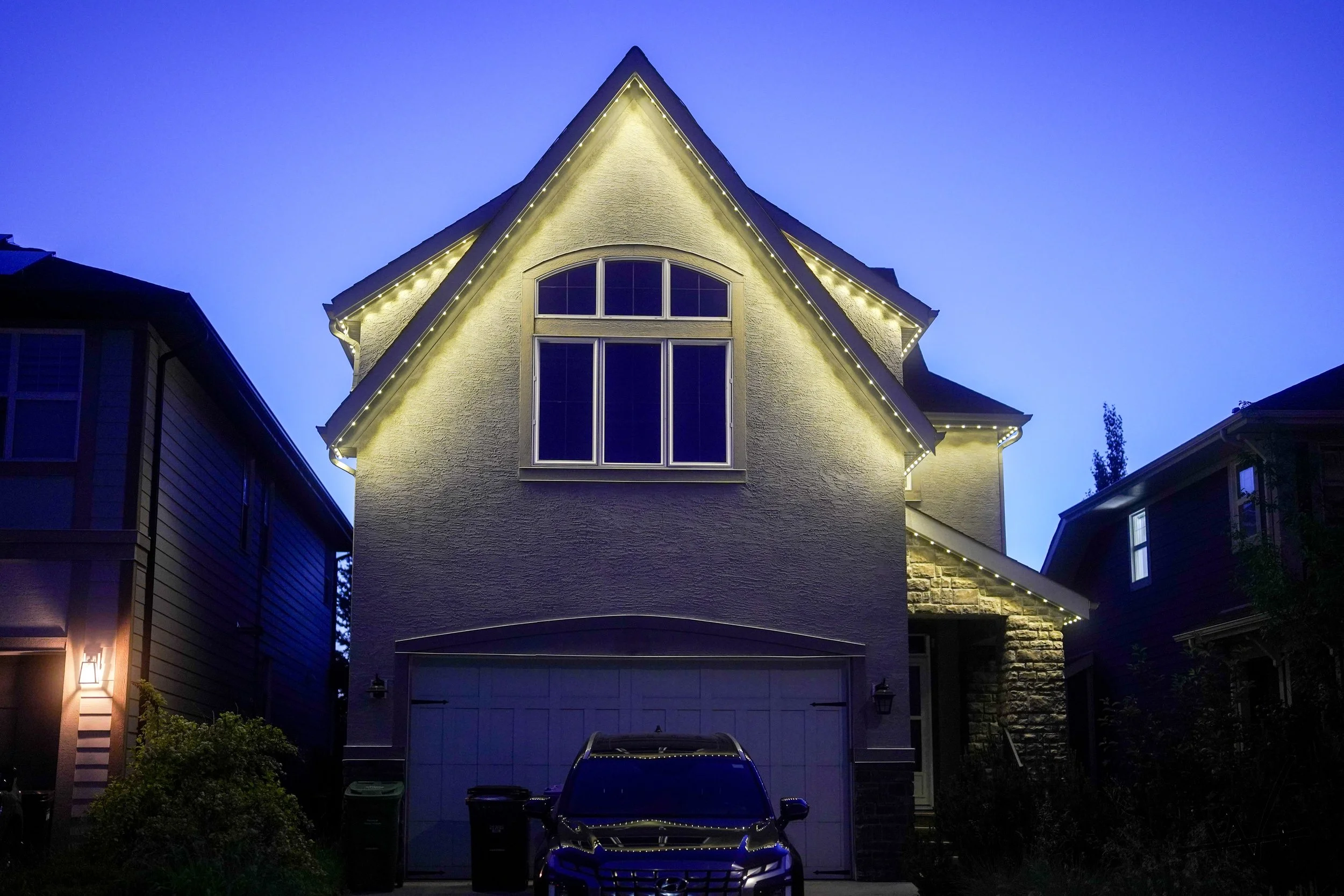 A two-story house illuminated at night with permanent track lighting along the roof edge, a large front window, a garage door, a car parked in front, and neighboring houses on each side.