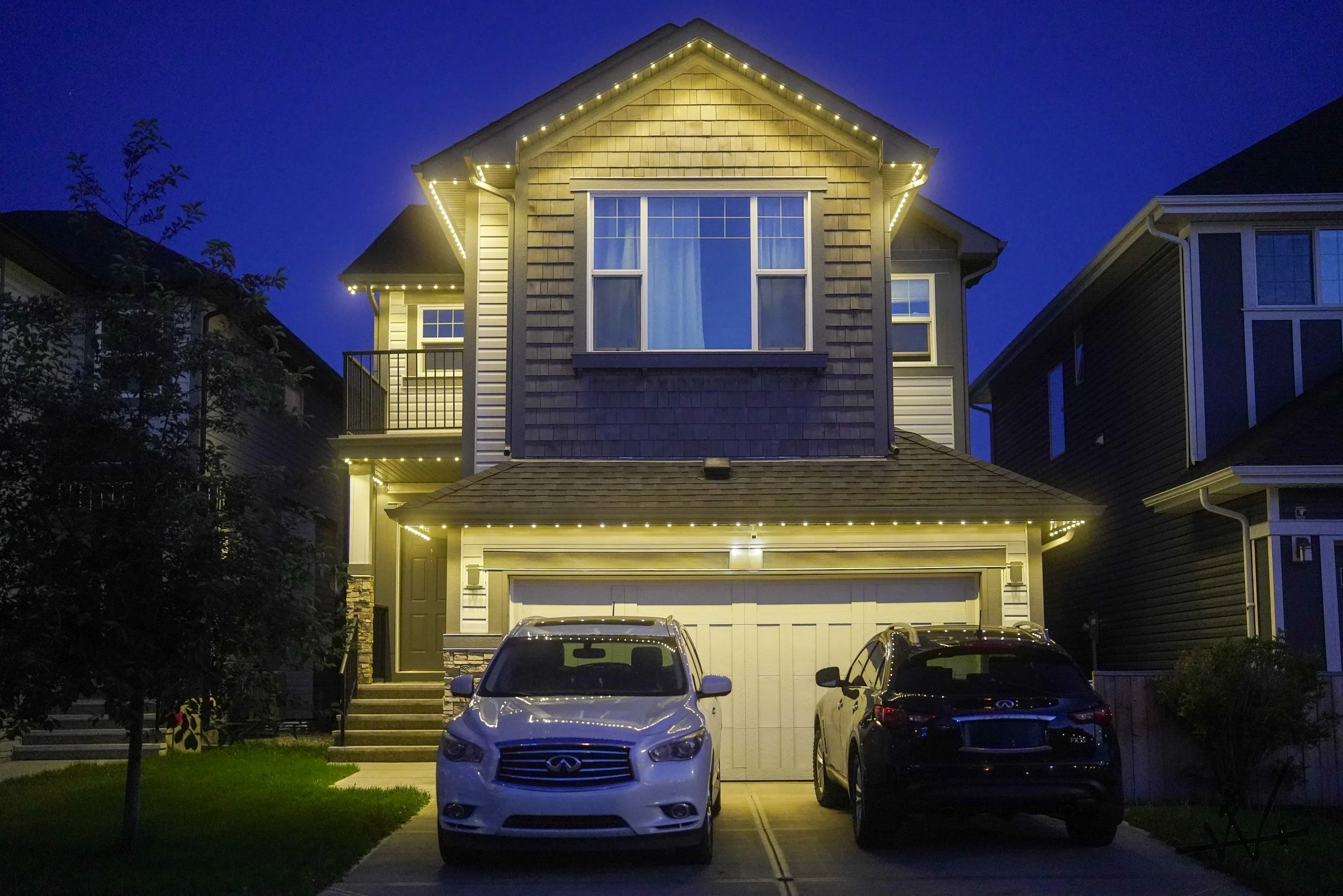 A modern, two-story house illuminated with permanent track lighting at night with two parked cars in the driveway.