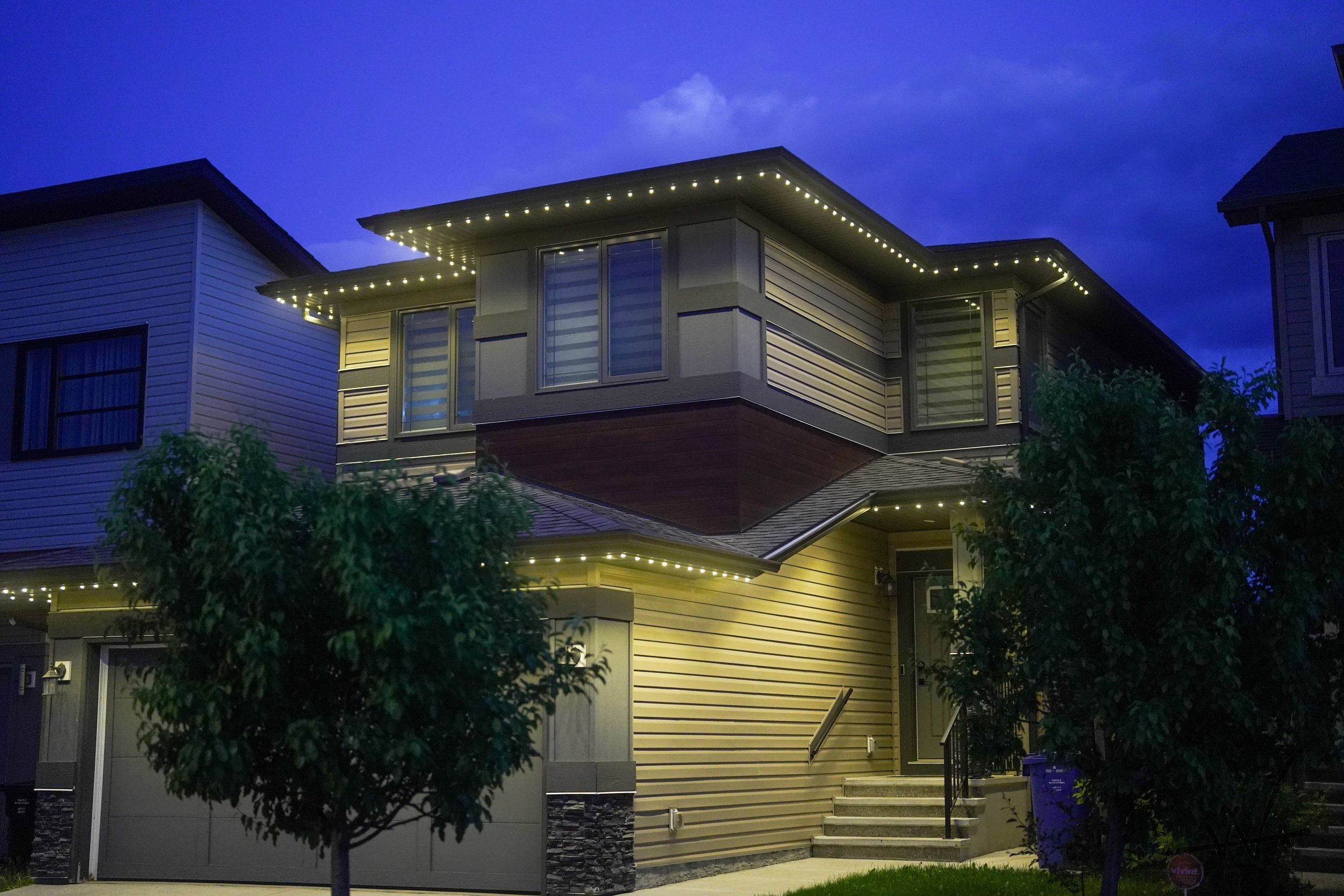 A modern two-story house with permanent track lighting at dusk, featuring horizontal siding, large windows, and a small front staircase.
