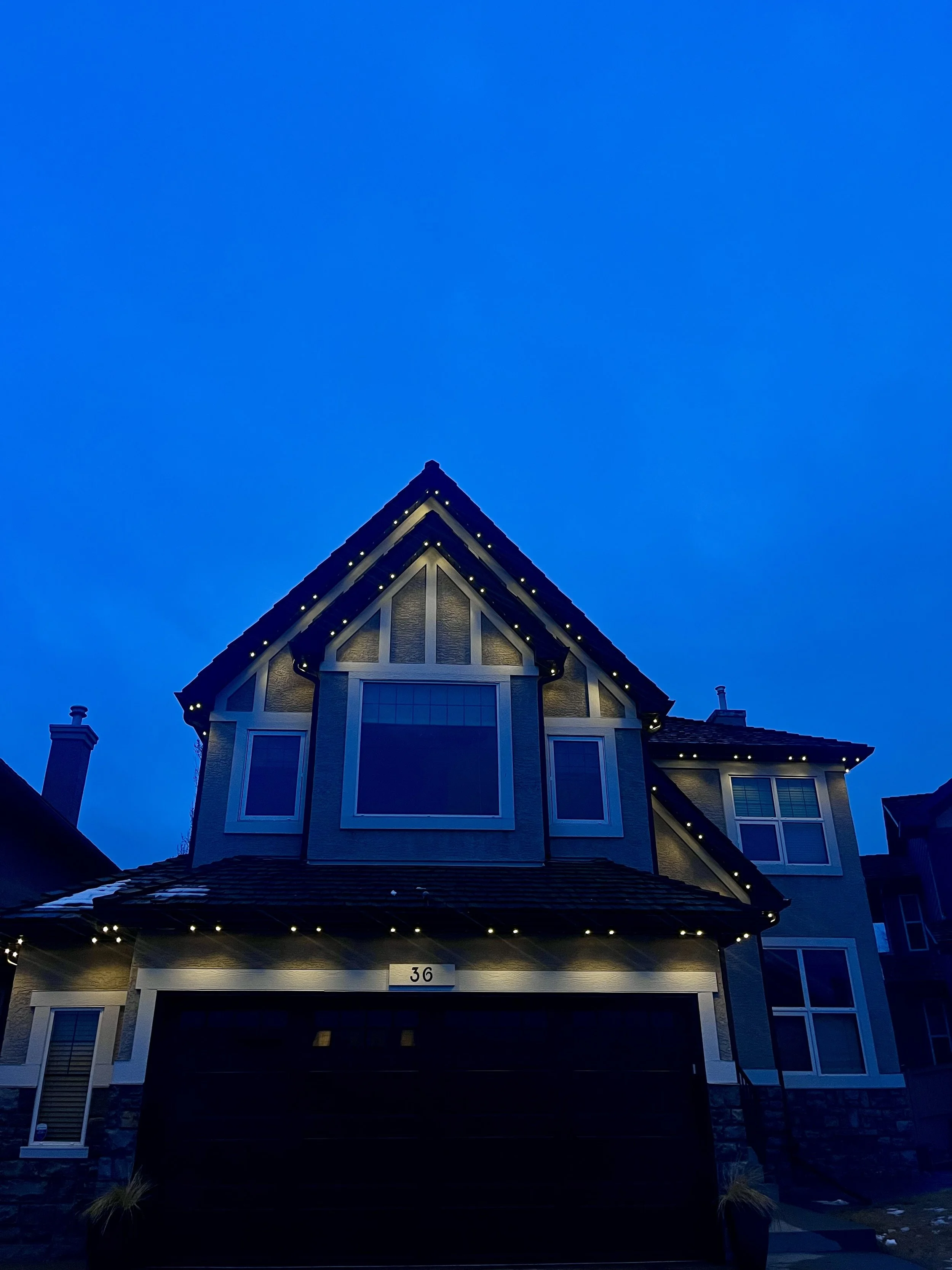 A large, modern house illuminated by permanent track lighting at dusk, with a prominent triangular roof and multiple windows, numbered 36.
