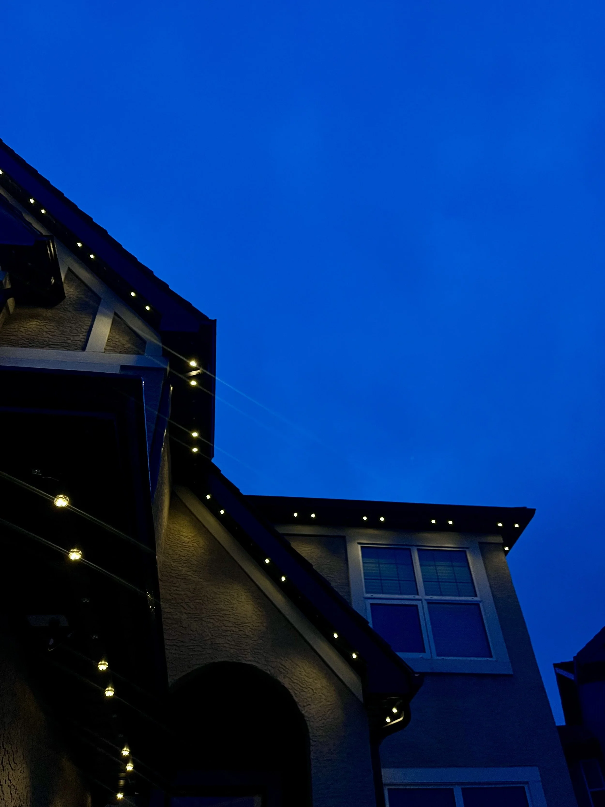 Nighttime view of a modern house with permanent track lighting along the roofline and window, under a dark blue sky.