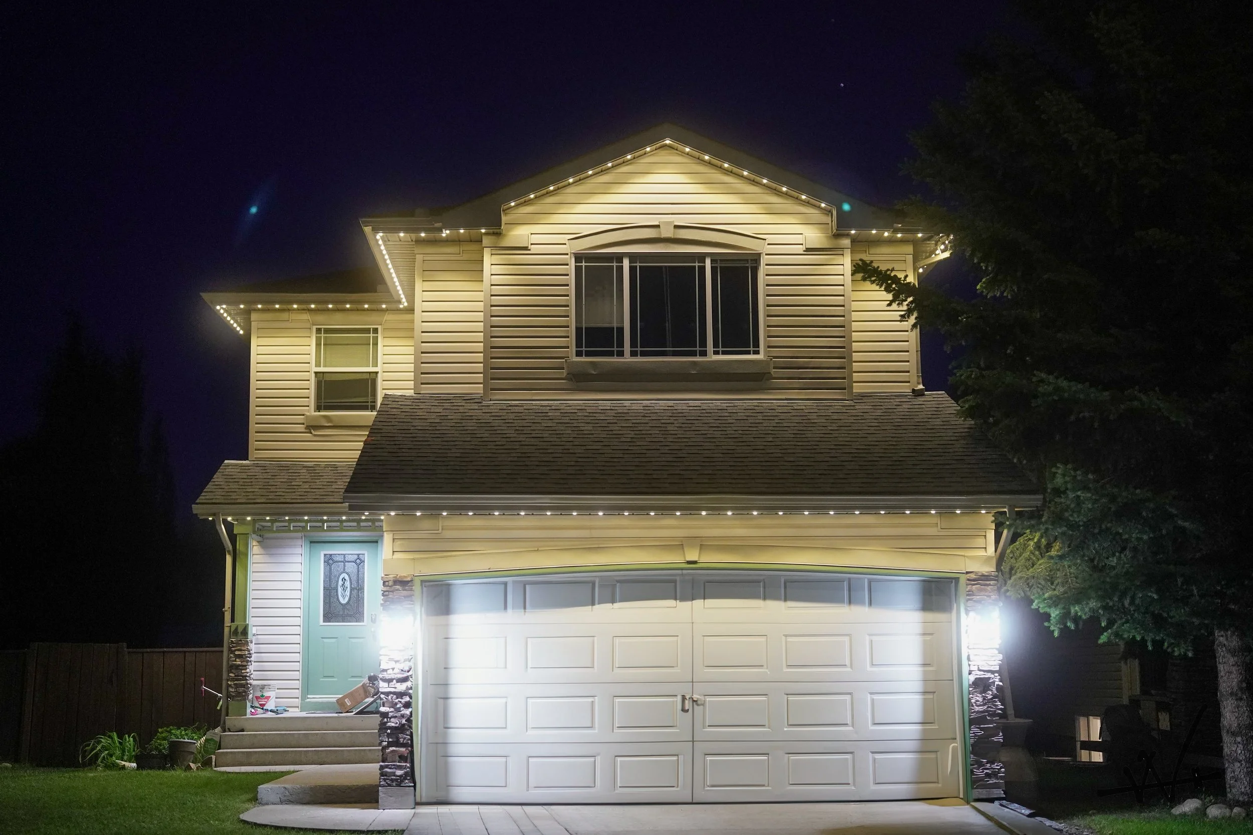 A two-story house at night with exterior lights outlining the roof and windows. The house has beige siding, a green front door, and a white garage door. A large tree is on the right side near the house, and a wooden fence is on the left. The sky is d