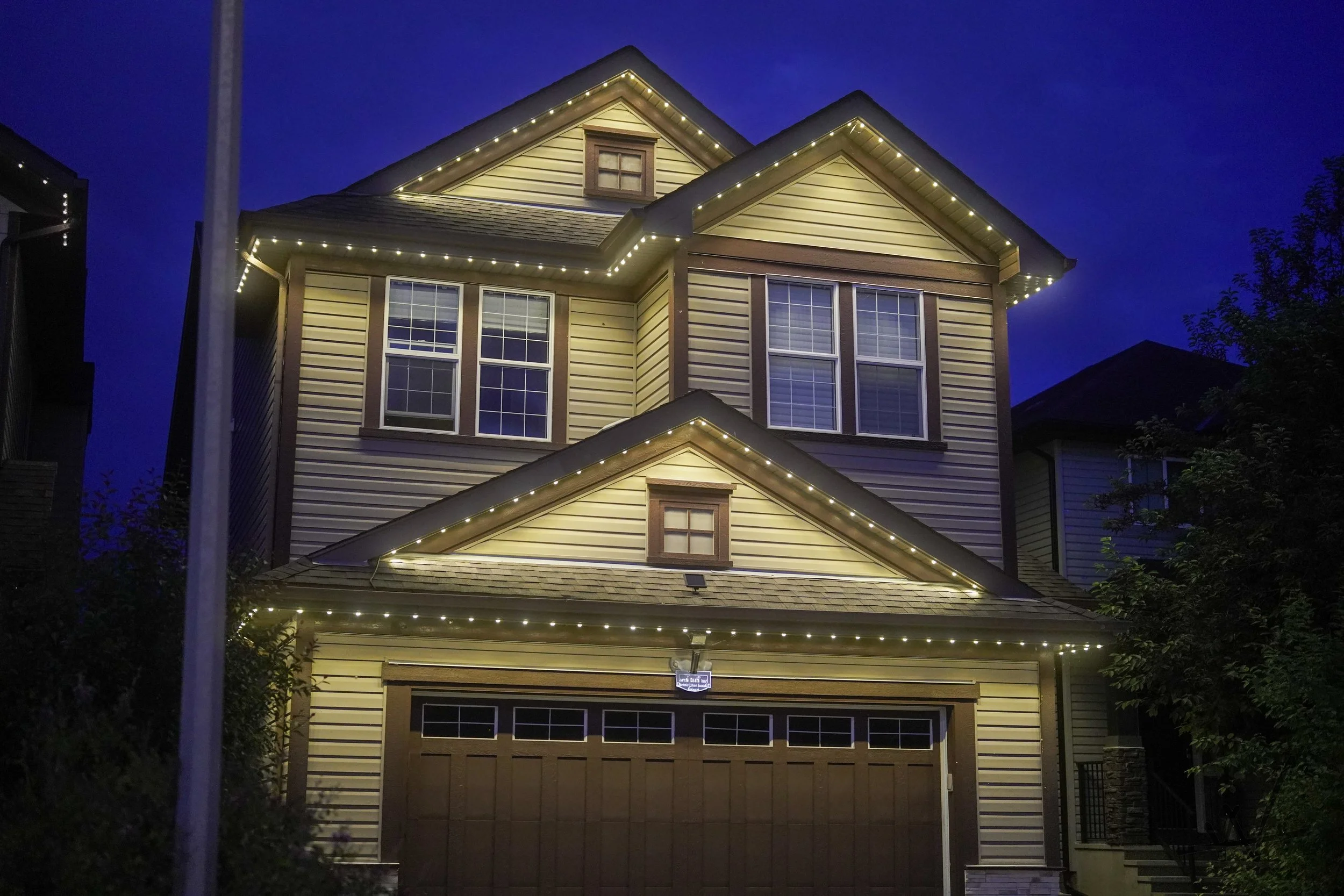 A multi-story house decorated with permanent track lighting along roof edges, illuminated at night, with a brown garage door and some trees nearby.