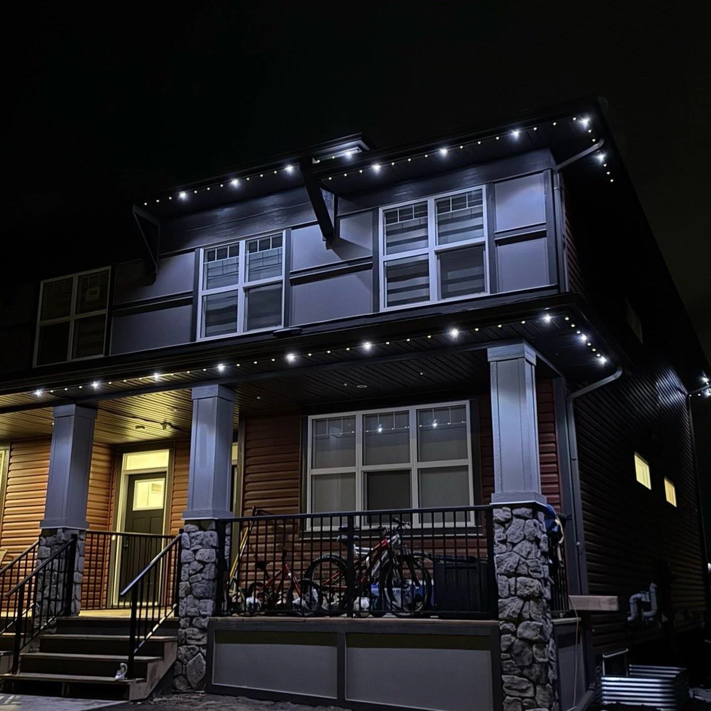 A modern two-story house at night with permanent track lighting, stone pillars, and bicycles on a porch.