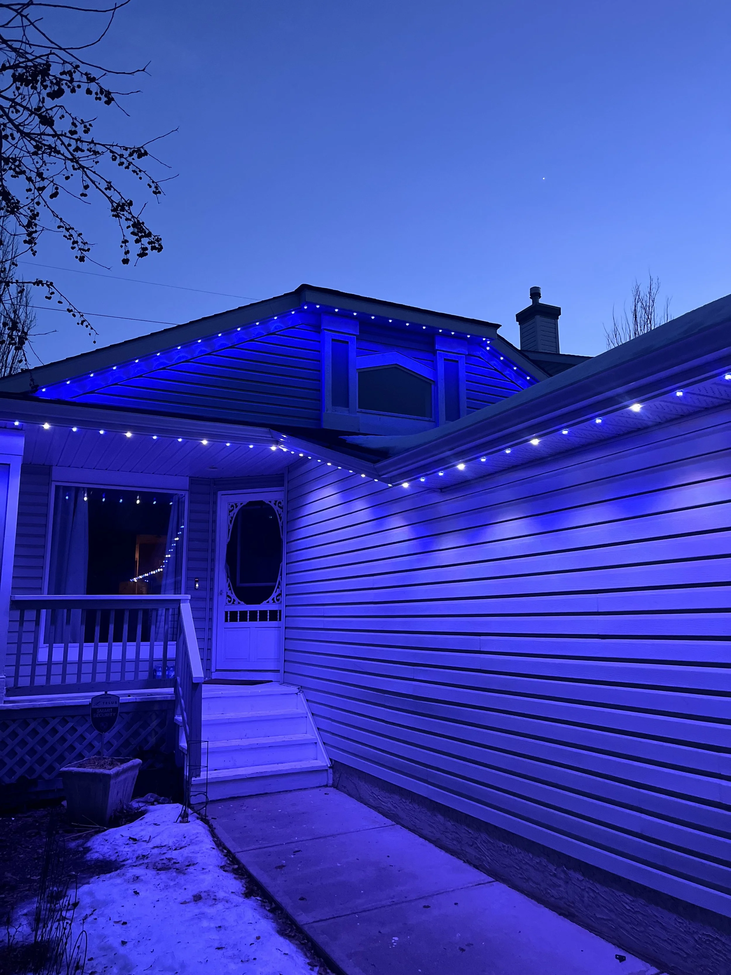 House exterior during dusk with permanent track lighting outlining the roof and porch, snow on the ground, and a tree branch in the upper left corner.