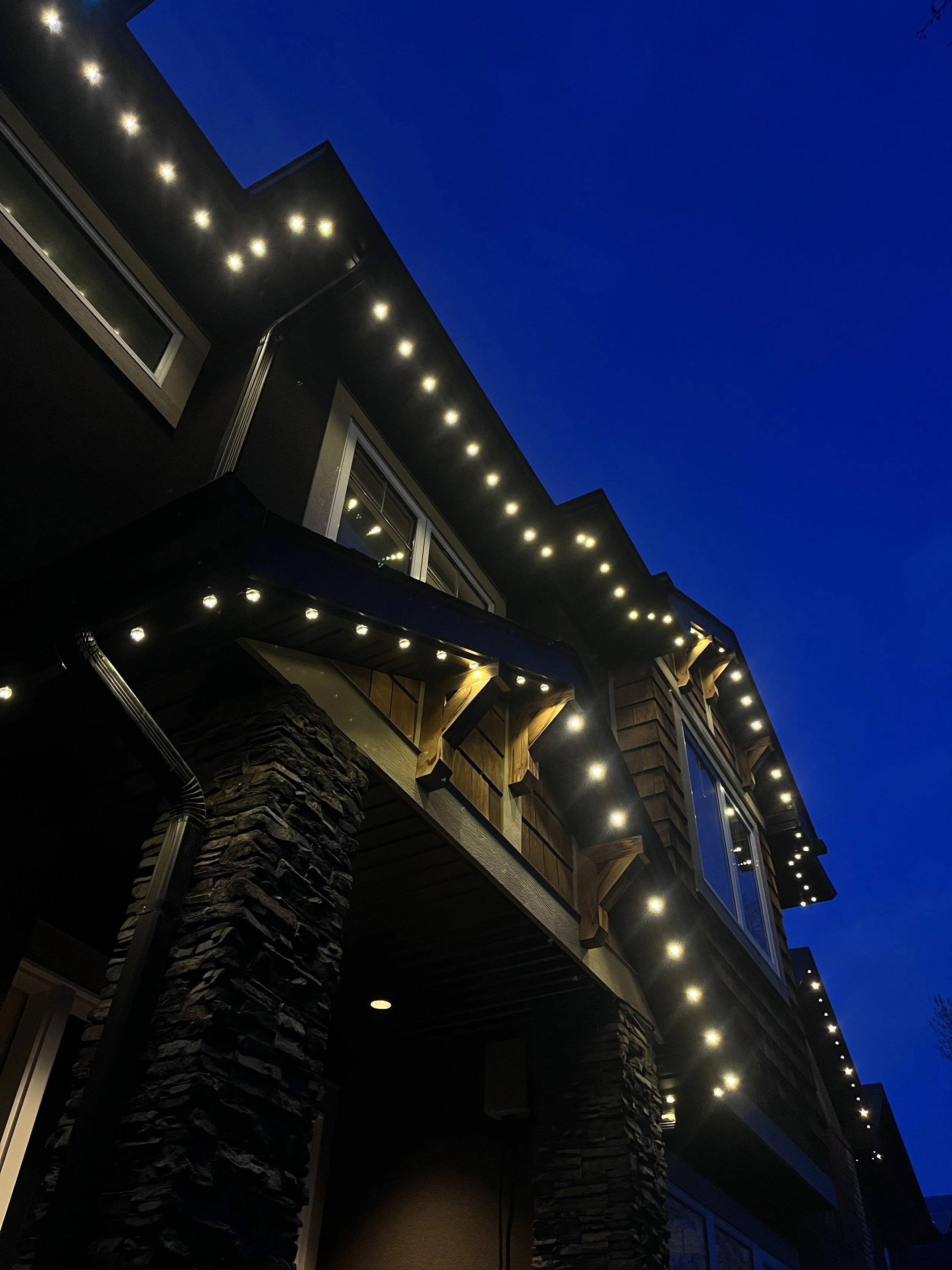 Exterior view of a modern house at night with permanent track lighting outlining the roof and balcony, dark stone pillars, and large windows.