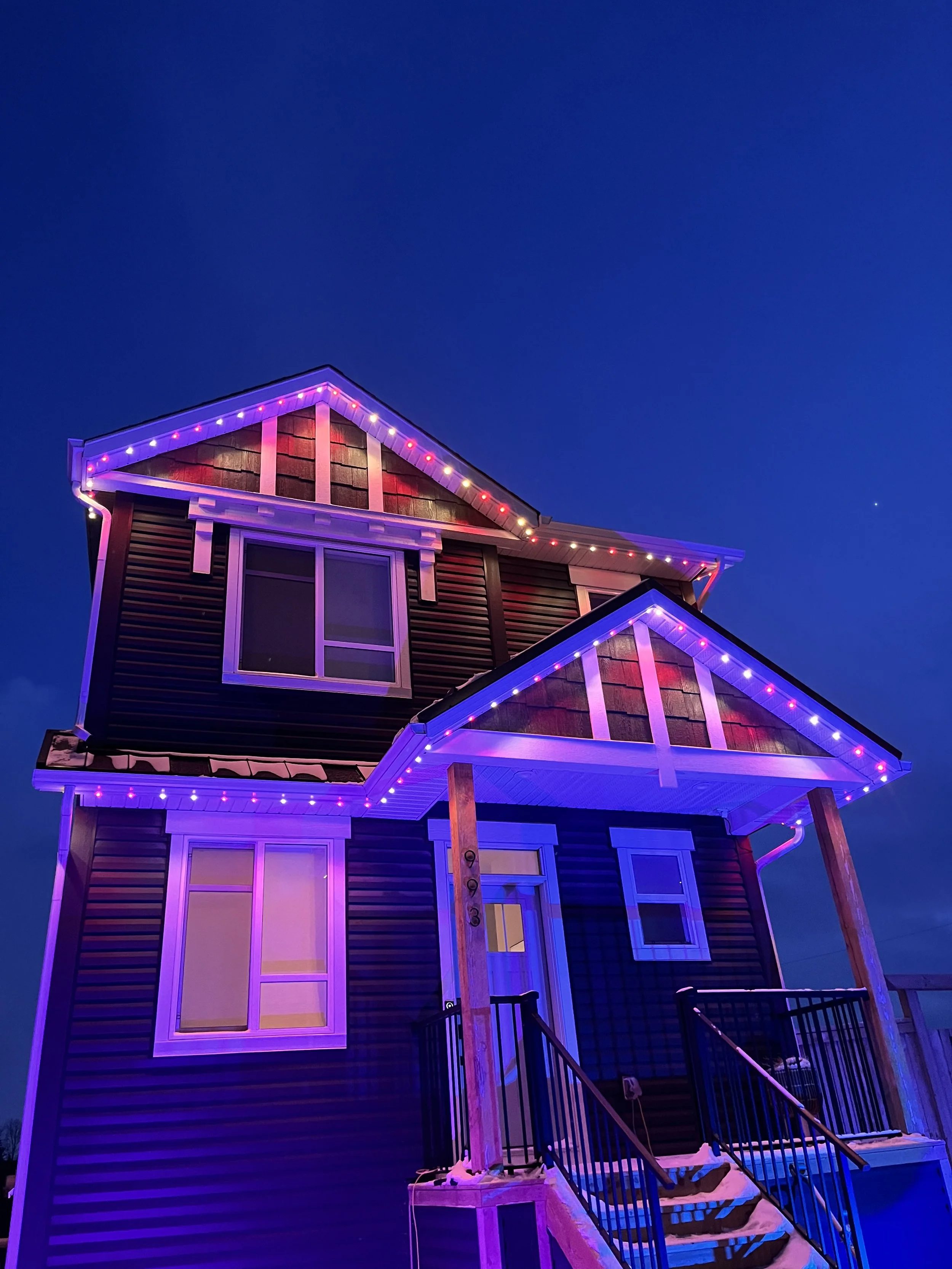 A two-story house decorated with permanent track lighting at dusk, with a blue sky in the background.