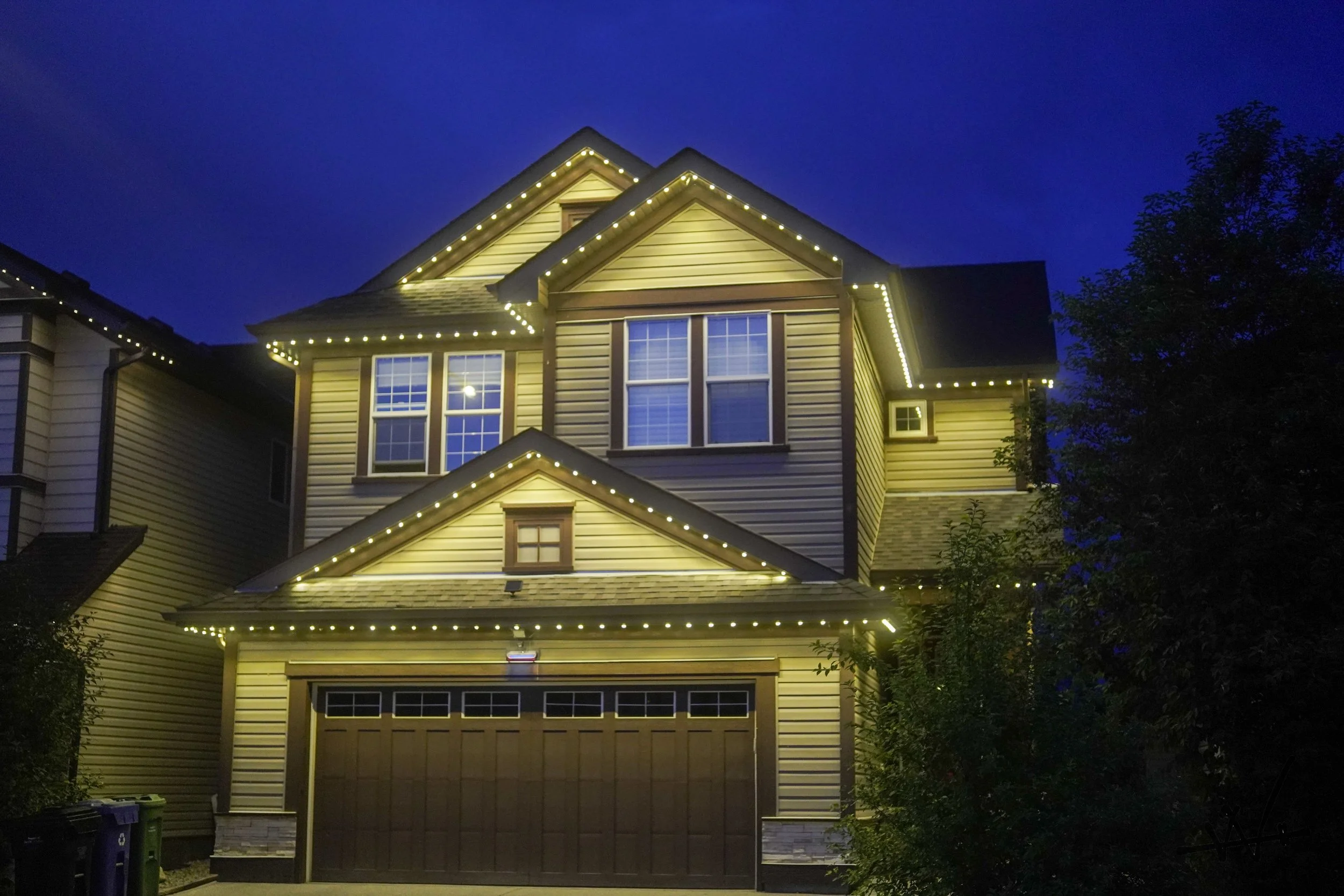 A two-story house decorated with permanent track lighting at night, with a brown garage door, beige siding, and a blue sky background.