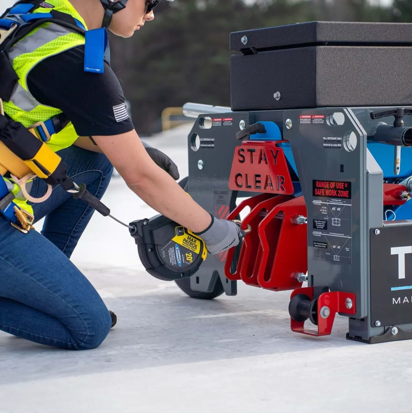 A person kneeling on snow, wearing safety gear, using a measuring tape to measure a piece of industrial equipment labeled "Stay Clear." The equipment is mounted on wheels and has warning labels and safety instructions. The person is focused on the task, wearing gloves, a safety vest, and protective glasses.