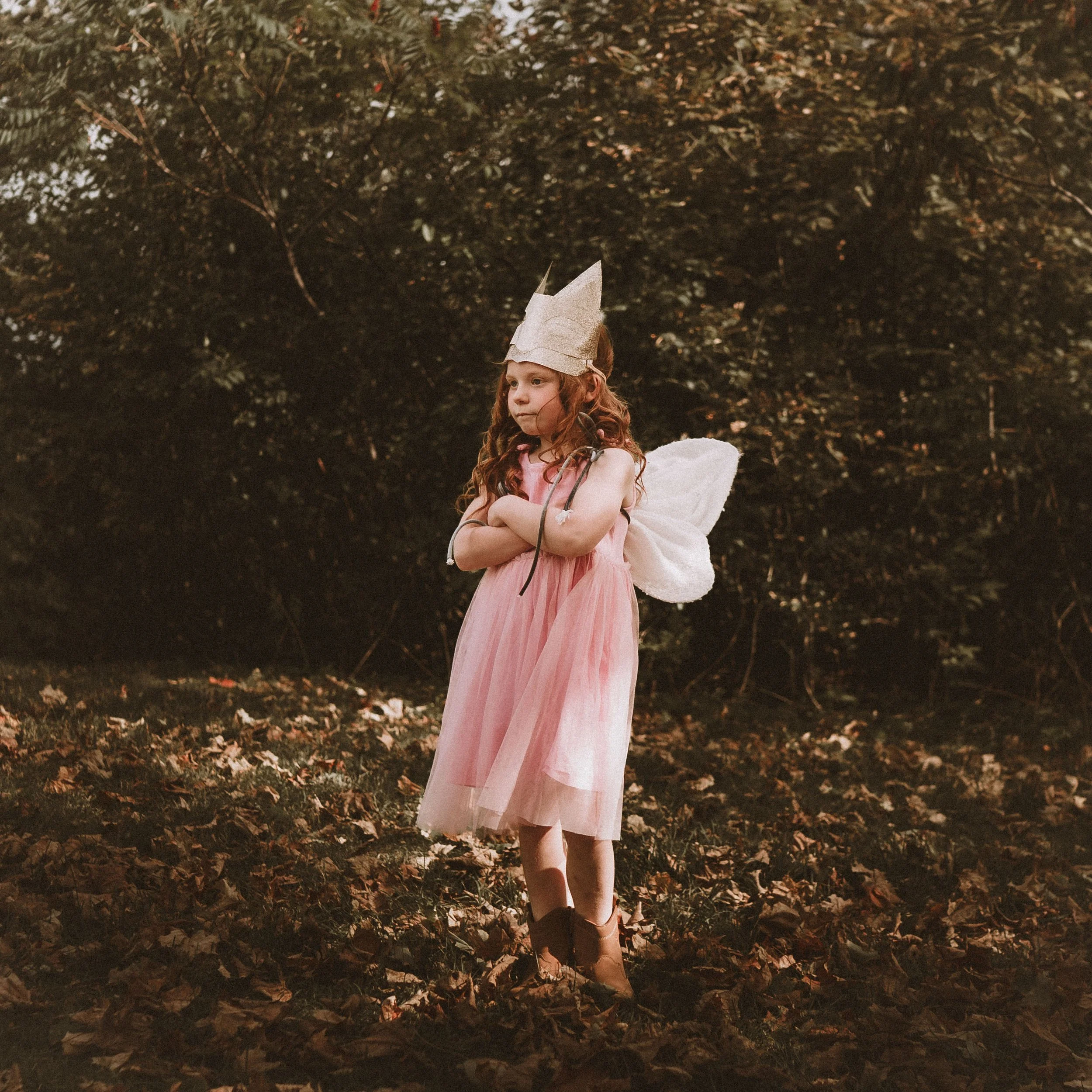 A young girl dressed as a fairy or princess, wearing a pink dress, fairy wings, a paper crown, and black boots, stands outdoors on a ground covered with leaves, with a background of trees.