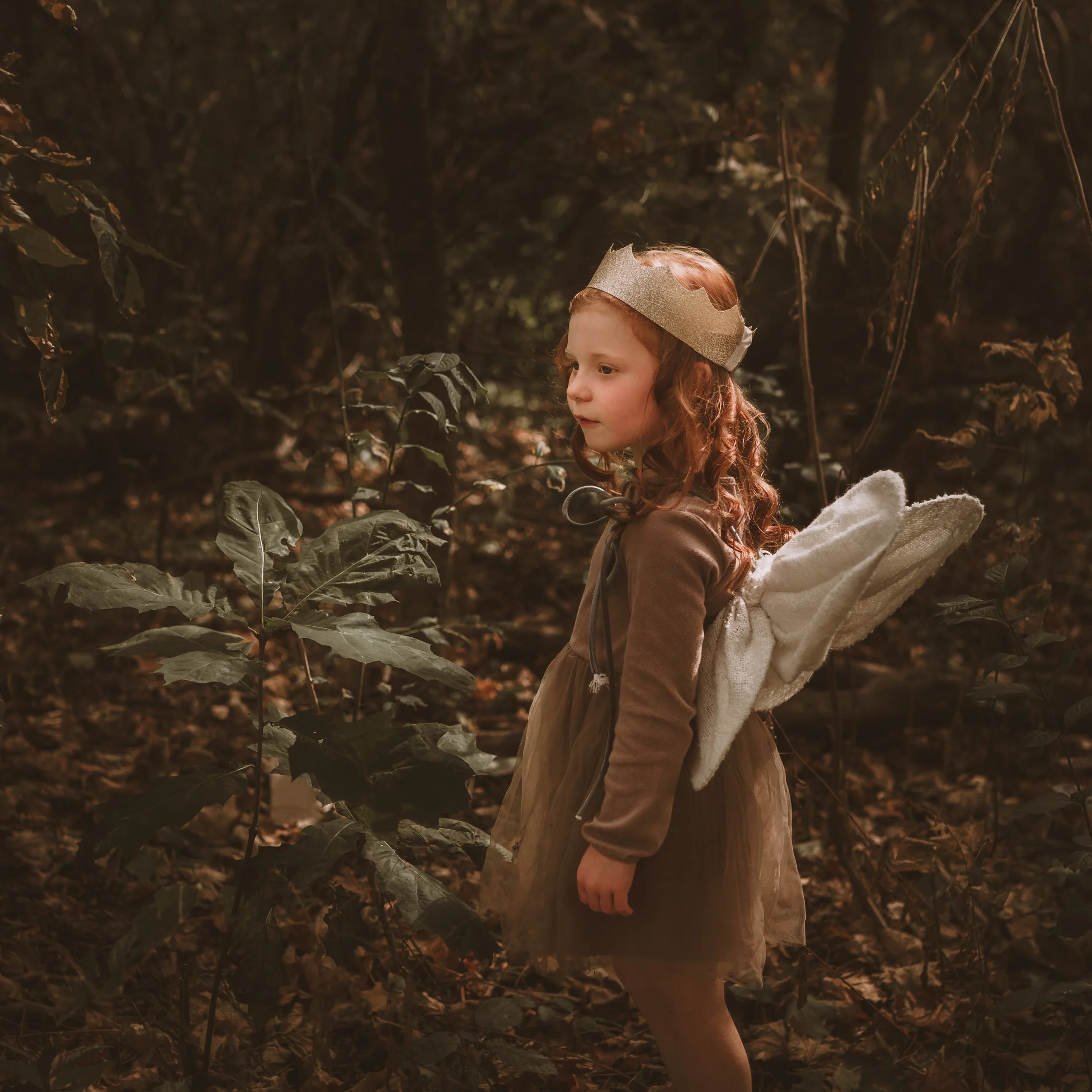 A child with red hair wearing a glittery paper crown and a beige jacket stands in a dark forest surrounded by fallen leaves and dense foliage.