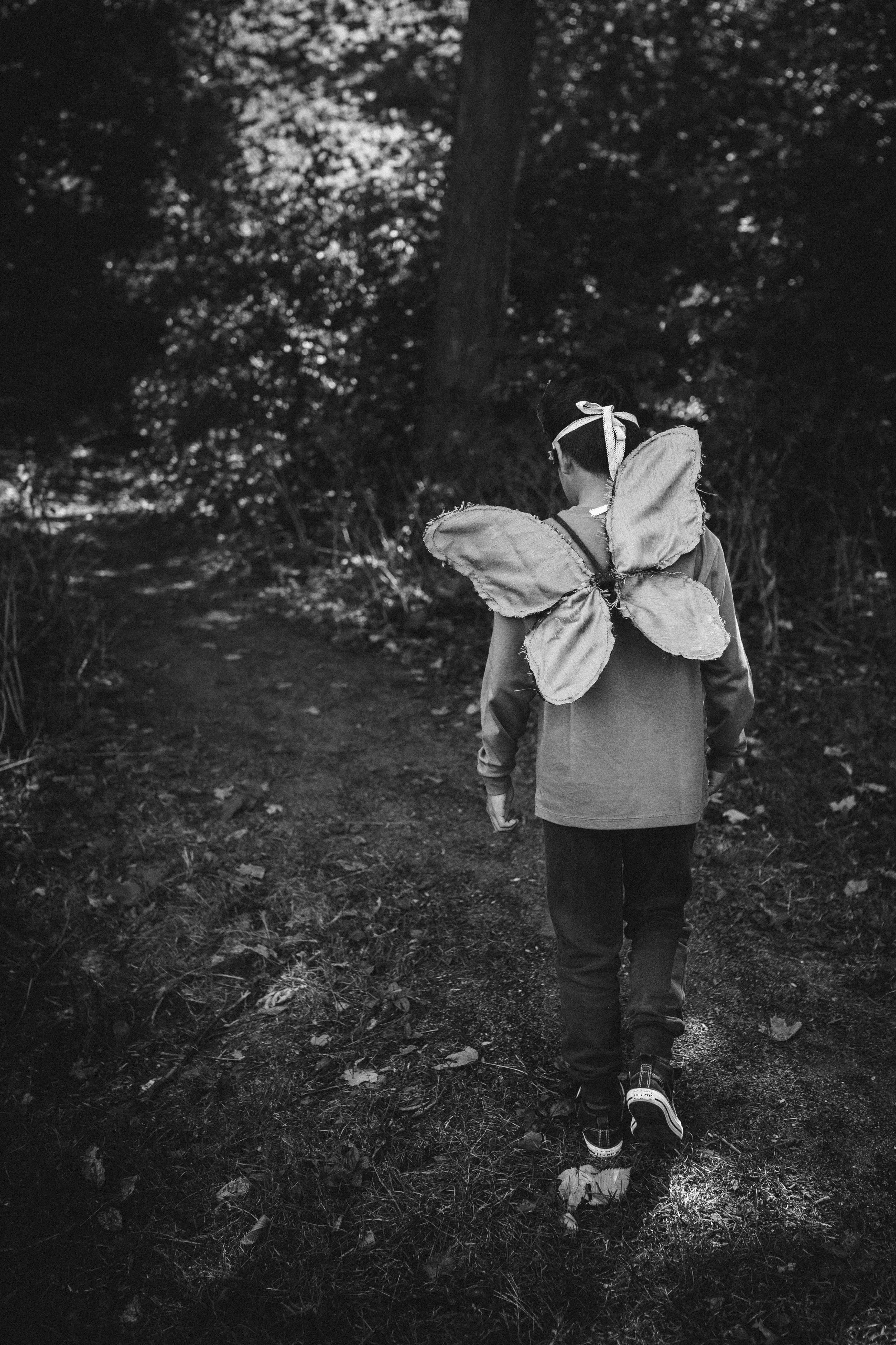 A black-and-white photo of a young boy walking on a dirt trail through a wooded area, wearing a backpack with fairy wings and a headband with ribbons.