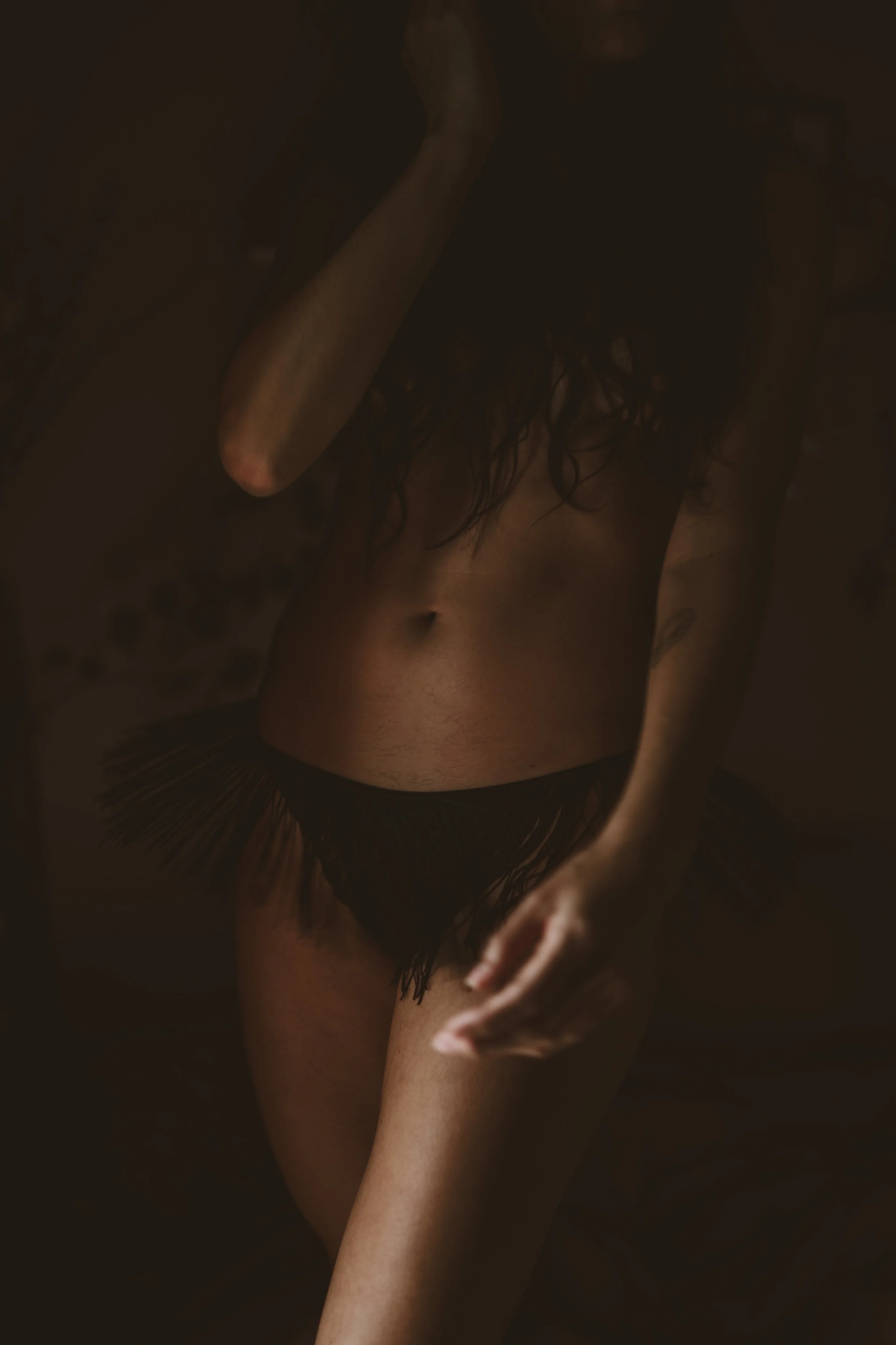 A person with long dark hair, wearing black fringed underwear, standing in a dark setting with moody lighting.